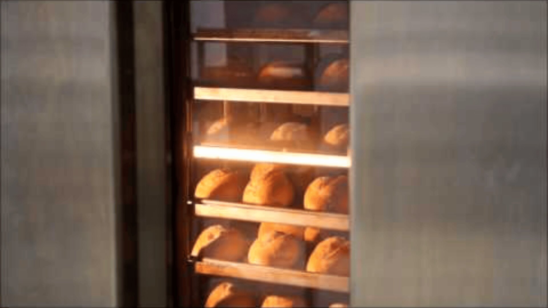 Rows of bread loaves baking inside an industrial oven, viewed through a glass door with metal framing and interior lighting illuminating the golden-brown crusts.