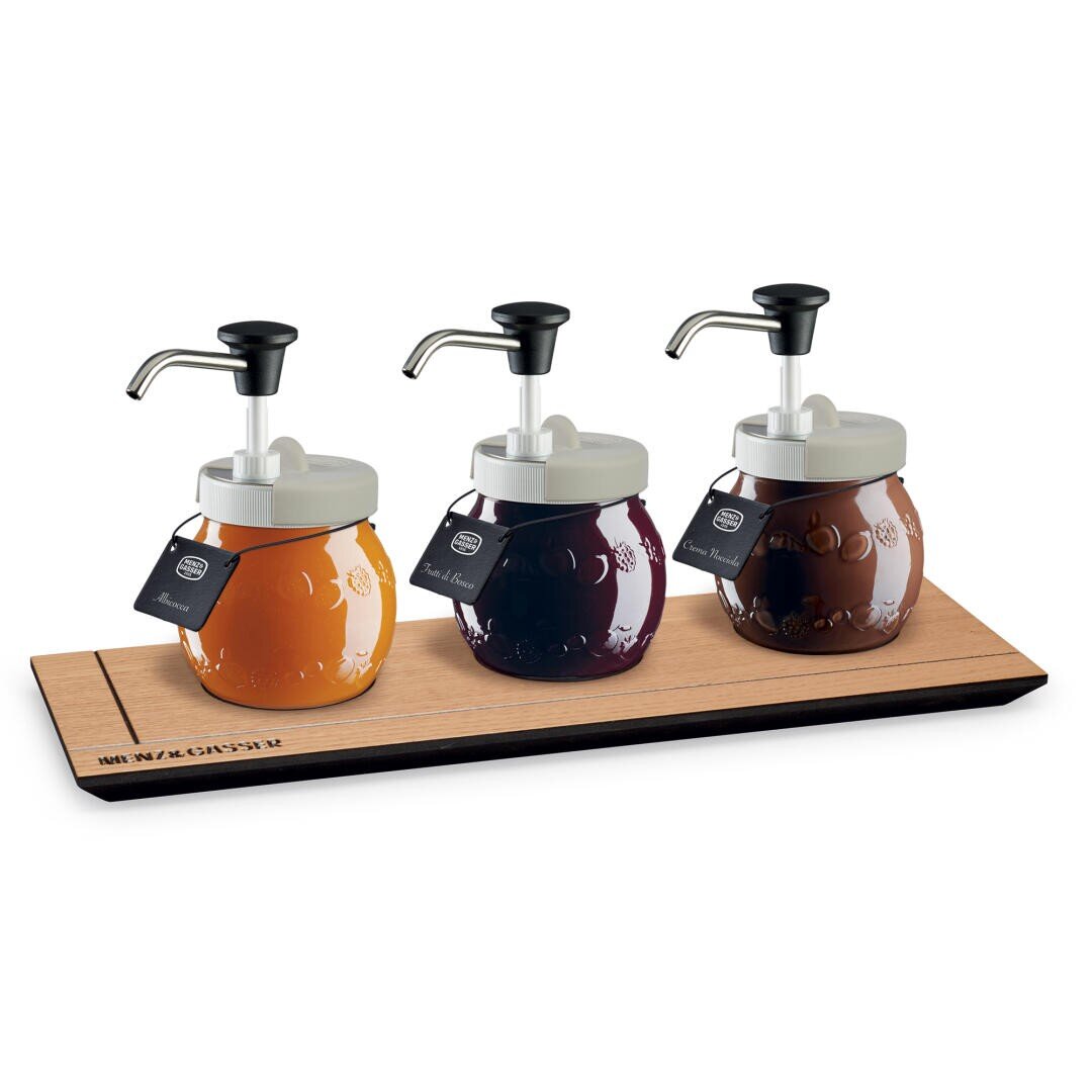 Drei gepumpte Gläser mit gelber, dunkelroter und brauner Soße sind auf einem Holztablett angeordnet. Jedes Glas hat einen weißen Deckel, einen Pumpspender und ein schwarzes Etikett, das am Hals befestigt ist.