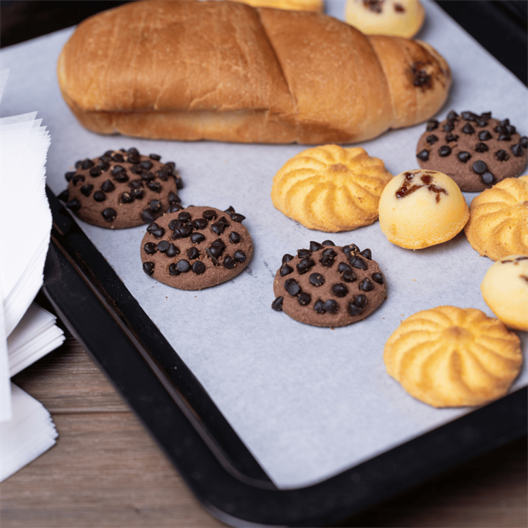 A baking tray with parchment paper holds chocolate chip cookies, yellow butter cookies, and a loaf of bread, with a stack of white napkins on the side.