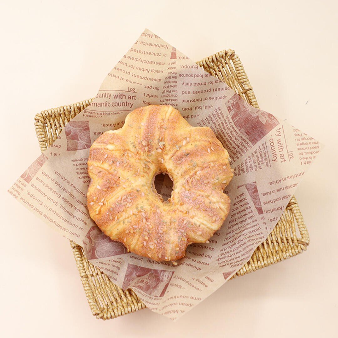 A round, rustic bread with a hole in the center sits on a square wicker basket, lined with brown printed parchment paper, against a light beige background.
