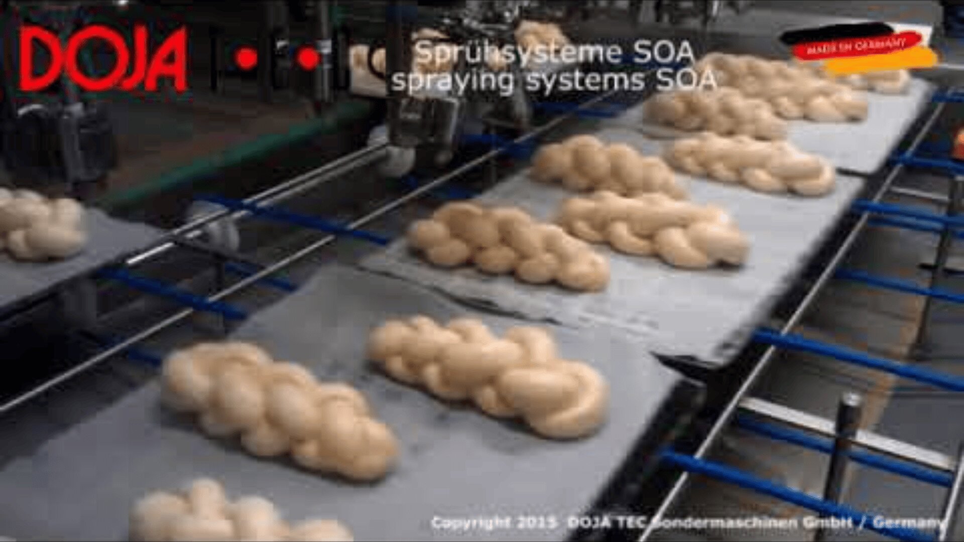 Baked goods move on a conveyor belt under industrial spraying systems labeled “Sprühsyteme SOA spraying systems SOA.” The DOJA logo and “Made in Germany” label appear in the image.