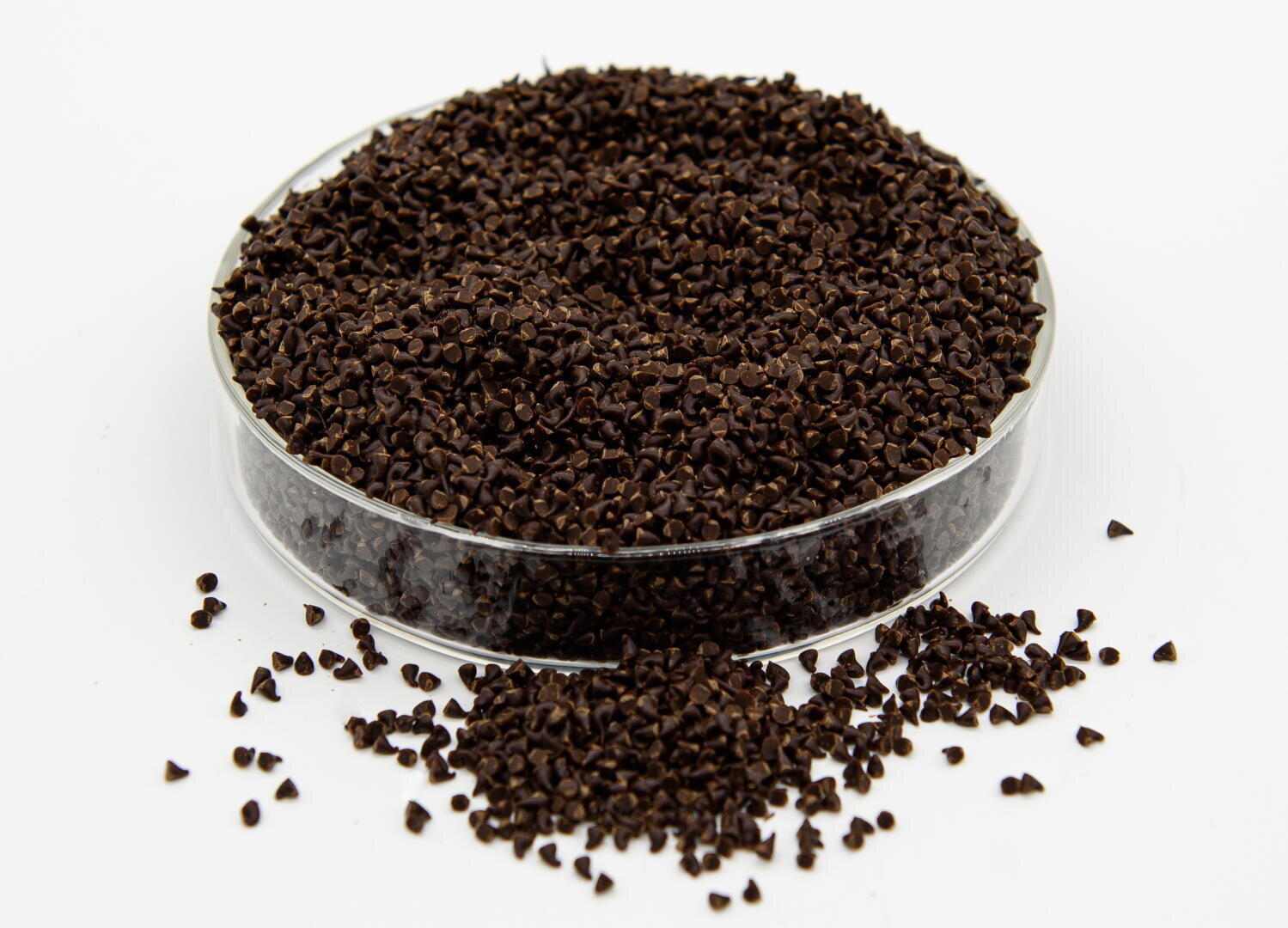 A clear dish filled with small, dark brown granules, with some granules scattered on the white surface around it.