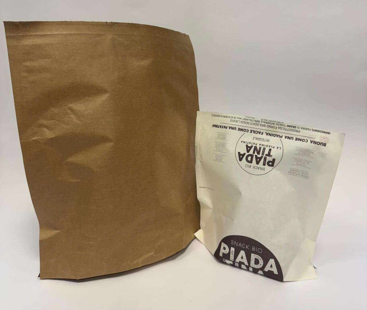 Two paper bags on a white surface: one large, plain brown bag on the left, and a smaller, white bag with PIADA LINDA and SNACK BIO printed on it on the right.