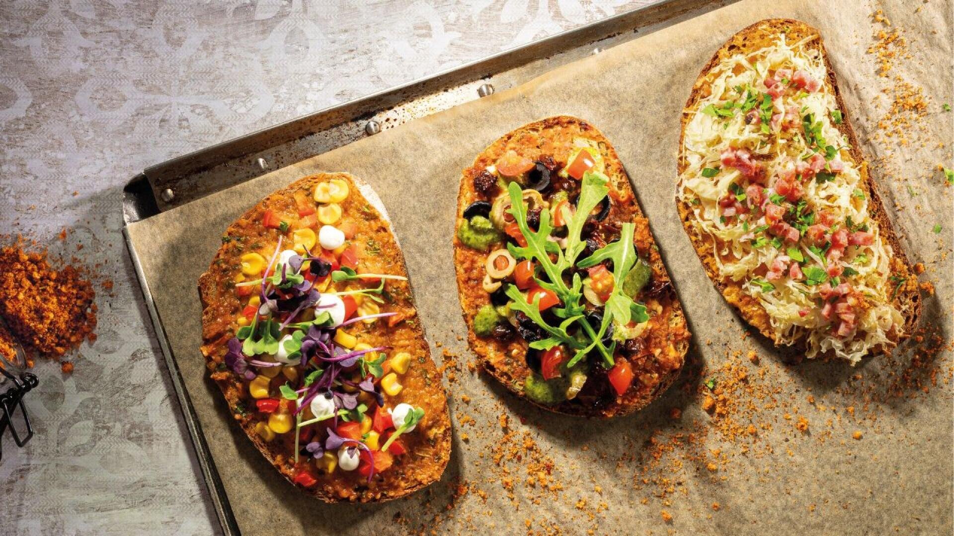 Drei mit buntem Gemüse, Grünzeug und Käse belegte Fladenbrote liegen auf Backpapier auf einem Backblech; jedes Fladenbrot weist eine andere Kombination frischer Zutaten auf.