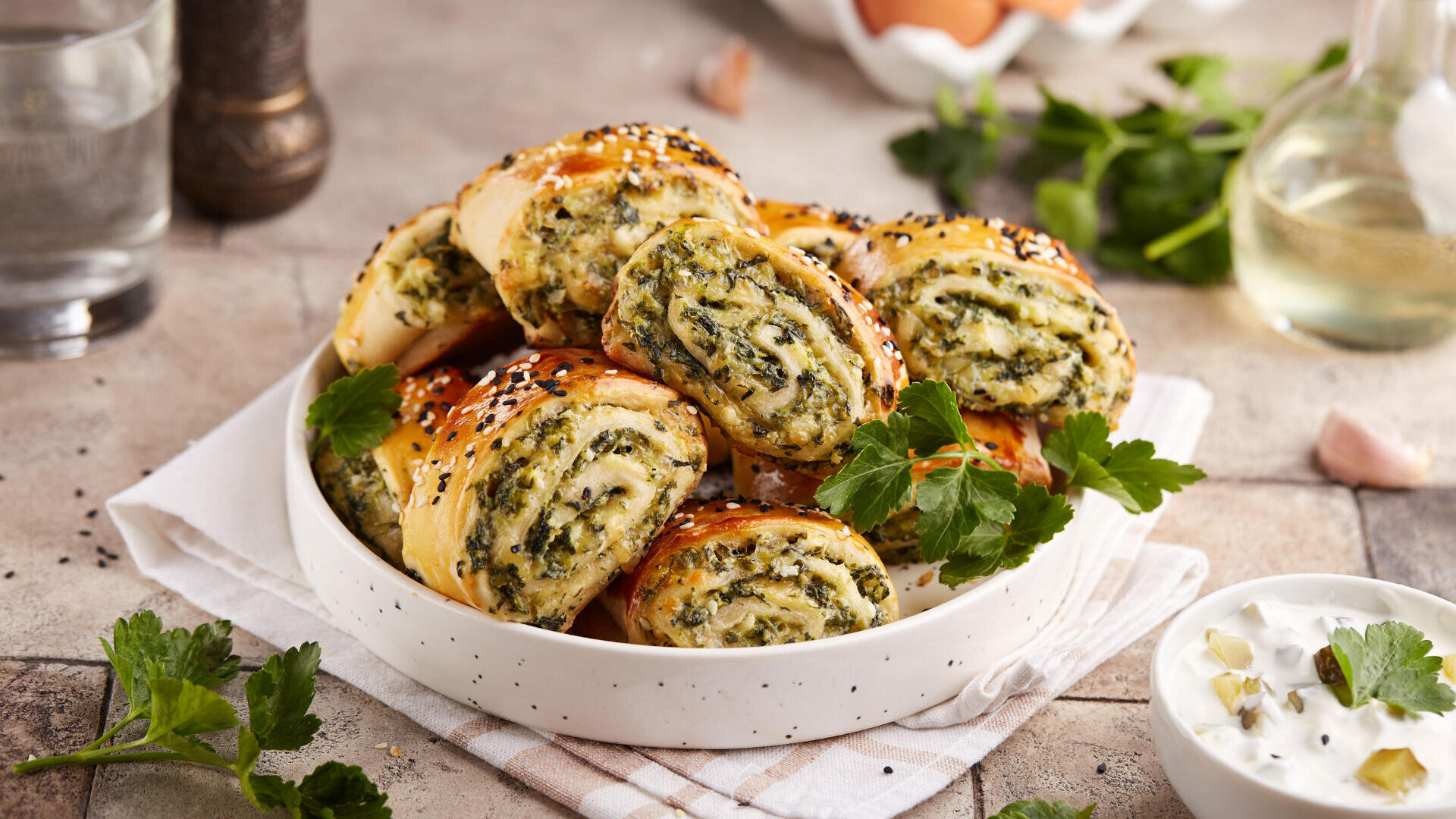 A bowl filled with slices of savory pastry rolls stuffed with spinach and cheese, garnished with sesame seeds, sits on a cloth napkin. Fresh parsley and a bowl of creamy dip are nearby.