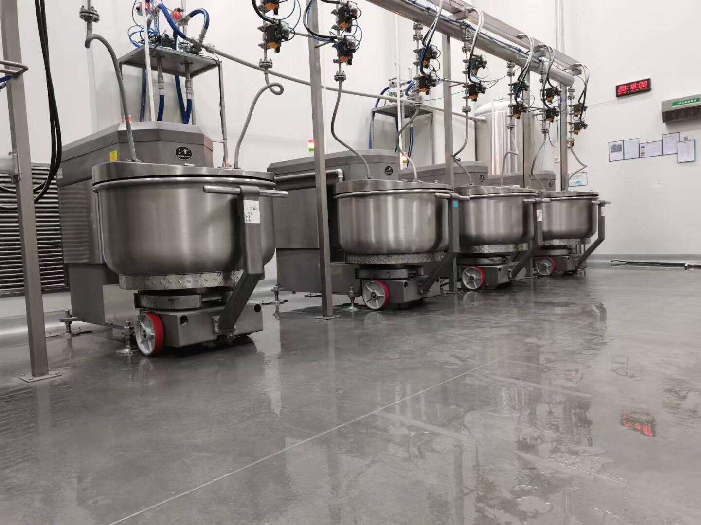A row of large industrial mixers with stainless steel bowls is lined up in a clean, modern food processing facility with polished floors and overhead piping.