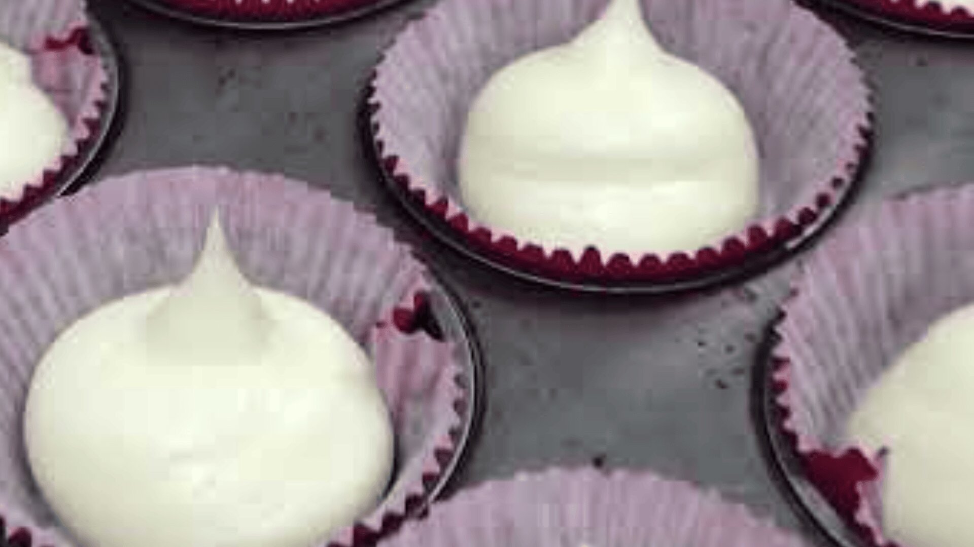 Close-up of raw white cupcake batter piped into pink paper cupcake liners arranged on a metal baking tray, ready to be baked.
