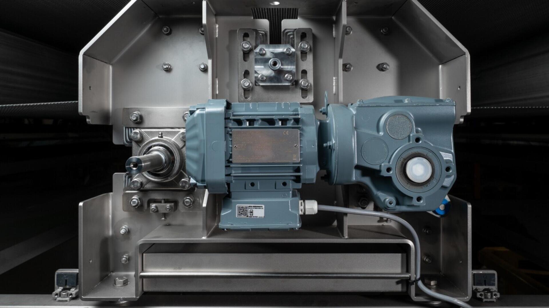 Close-up view of a mechanical gearbox and motor assembly mounted on a metal frame, featuring exposed gears, wiring, and various bolts in an industrial setting.