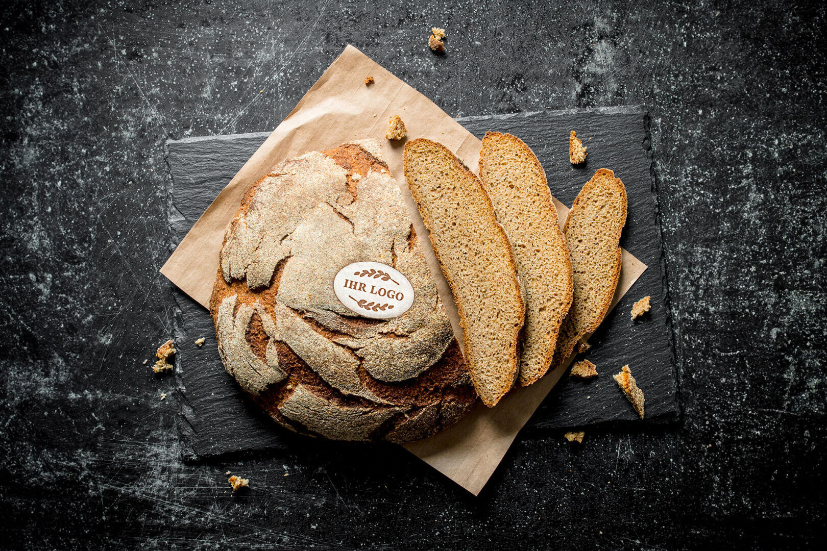 Runder, rustikaler Laib Brot mit dem Etikett „IHR LOGO“ auf der Oberseite, auf braunem Papier und einer dunklen Schiefertafel platziert, mit drei Brotscheiben und Krümeln auf einer strukturierten schwarzen Oberfläche.