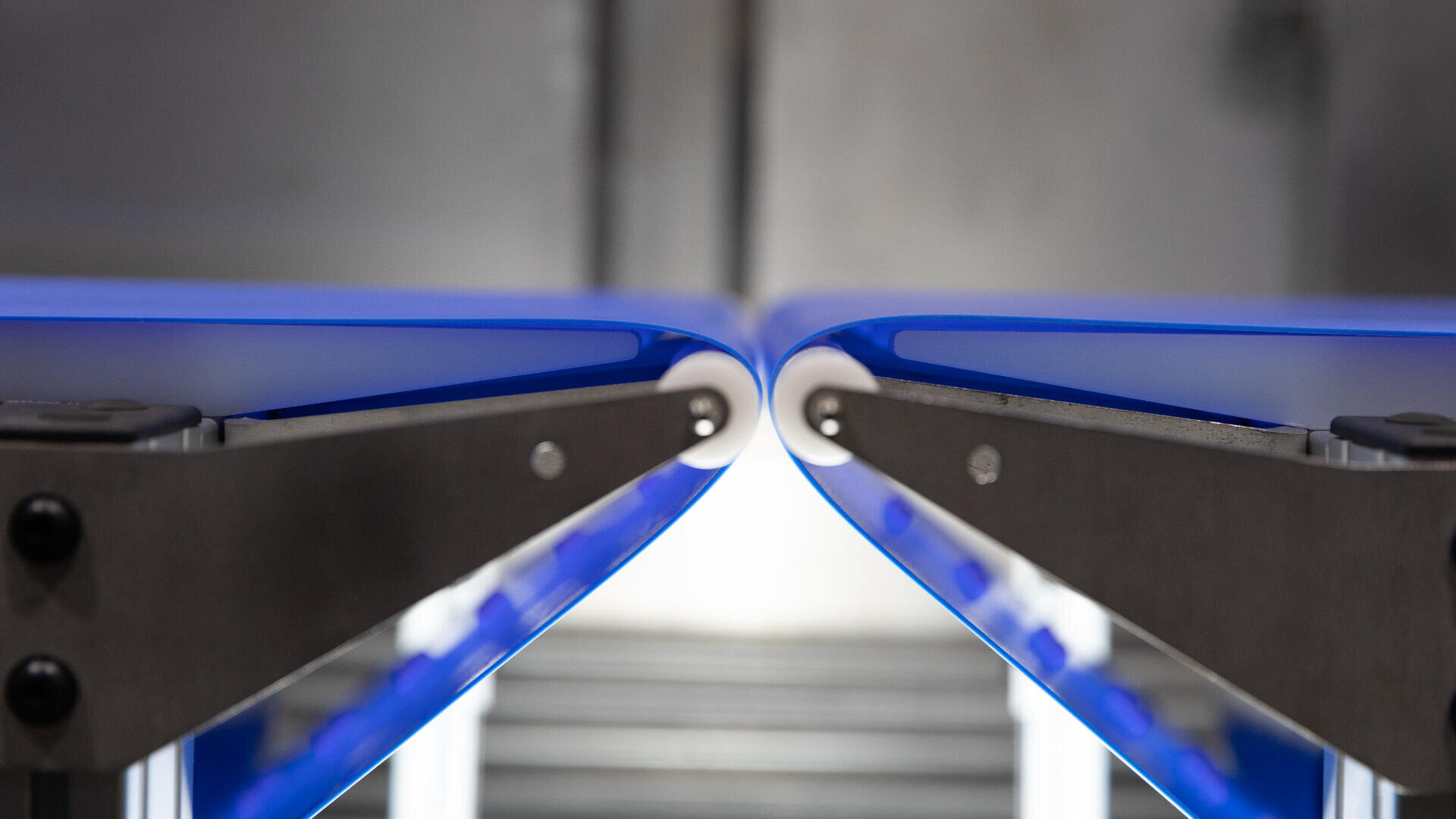 Close-up view of two blue conveyor belts coming together, with metal supports visible. The belts are aligned and facing each other, creating a narrow gap in the center. The background is blurred.