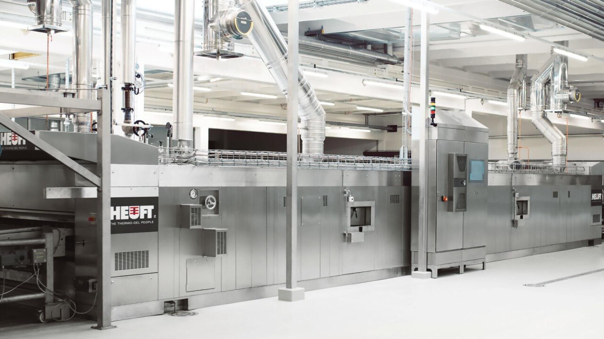 A large, industrial stainless steel food processing machine with vents and control panels stands in a bright, clean factory setting with overhead pipes and lighting. The machine is labeled HEUFT.