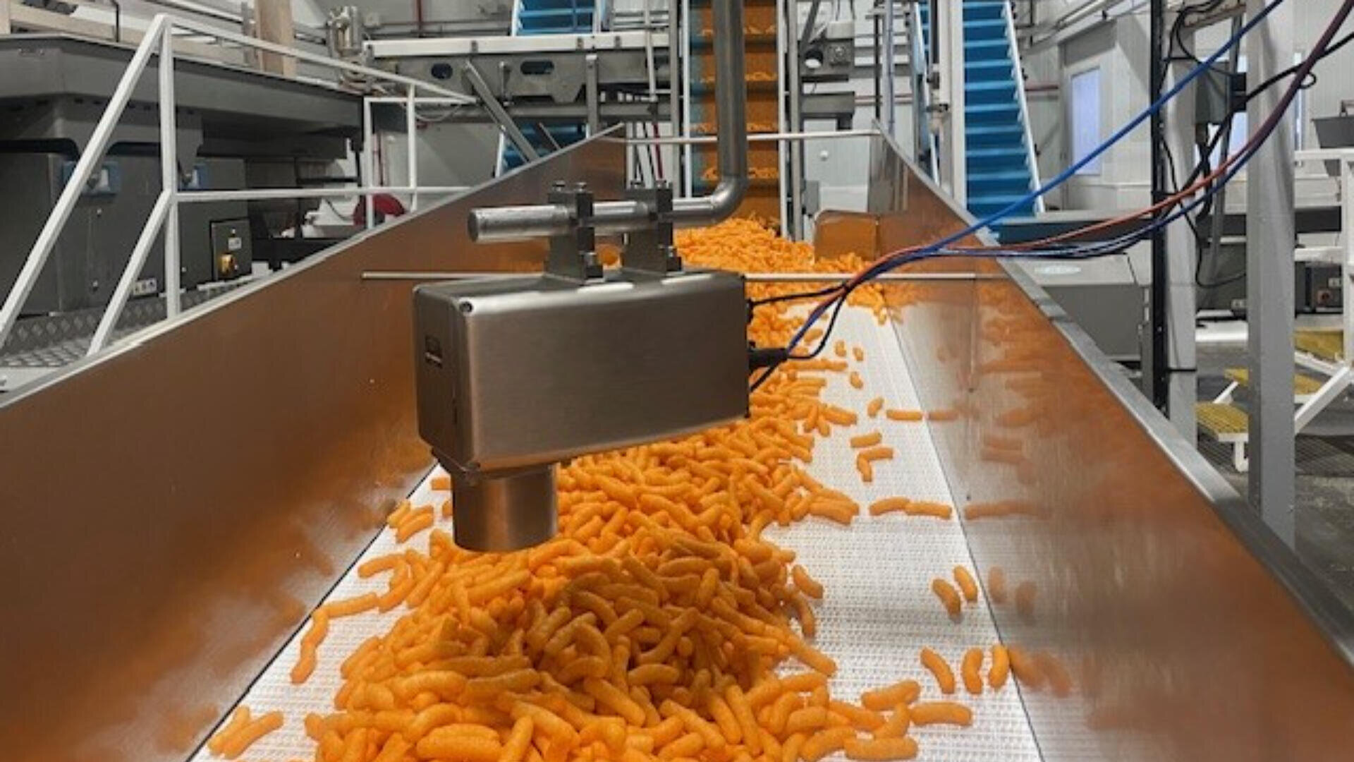 Orange cheese snacks are moving along a conveyor belt in a food processing plant, with a metal machine hovering above inspecting or distributing them. Equipment and machinery are visible in the background.