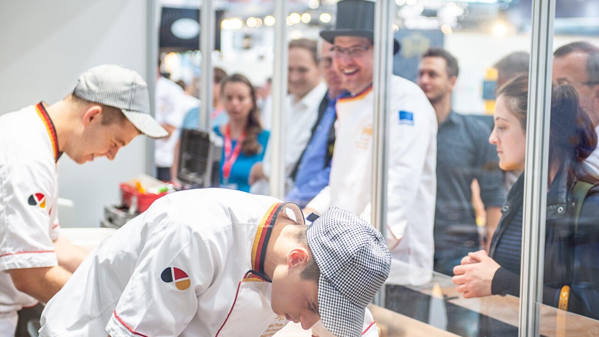 Two chefs in white uniforms work behind a glass partition while a group of people, some smiling, watch them from the other side. One chef is bending over his work, and the scene appears lively and focused.
