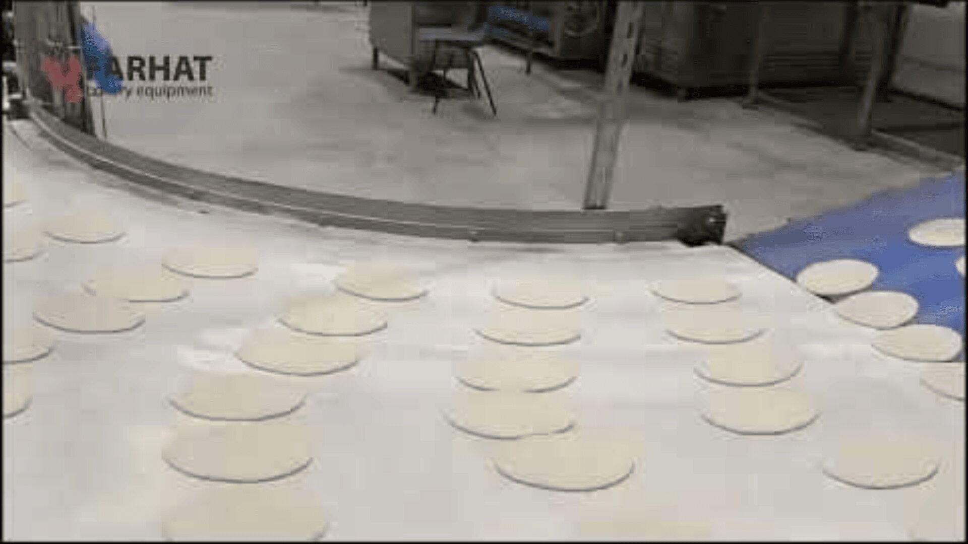 Conveyor belt with evenly spaced round pieces of dough in a factory setting, with machinery and equipment visible in the background.