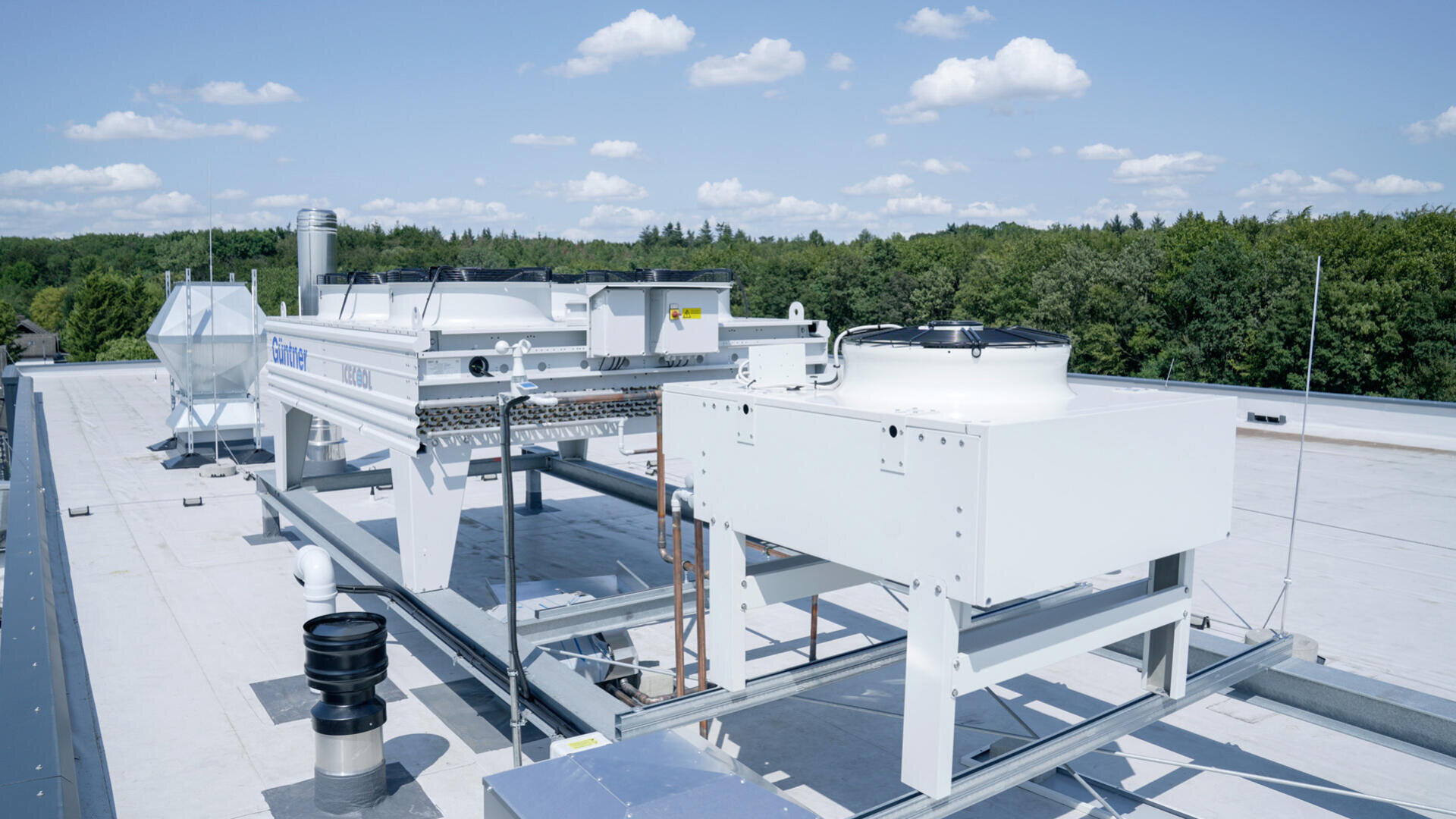 Industrial HVAC equipment and ventilation units installed on a flat rooftop, surrounded by a forest of green trees under a blue sky with scattered clouds.