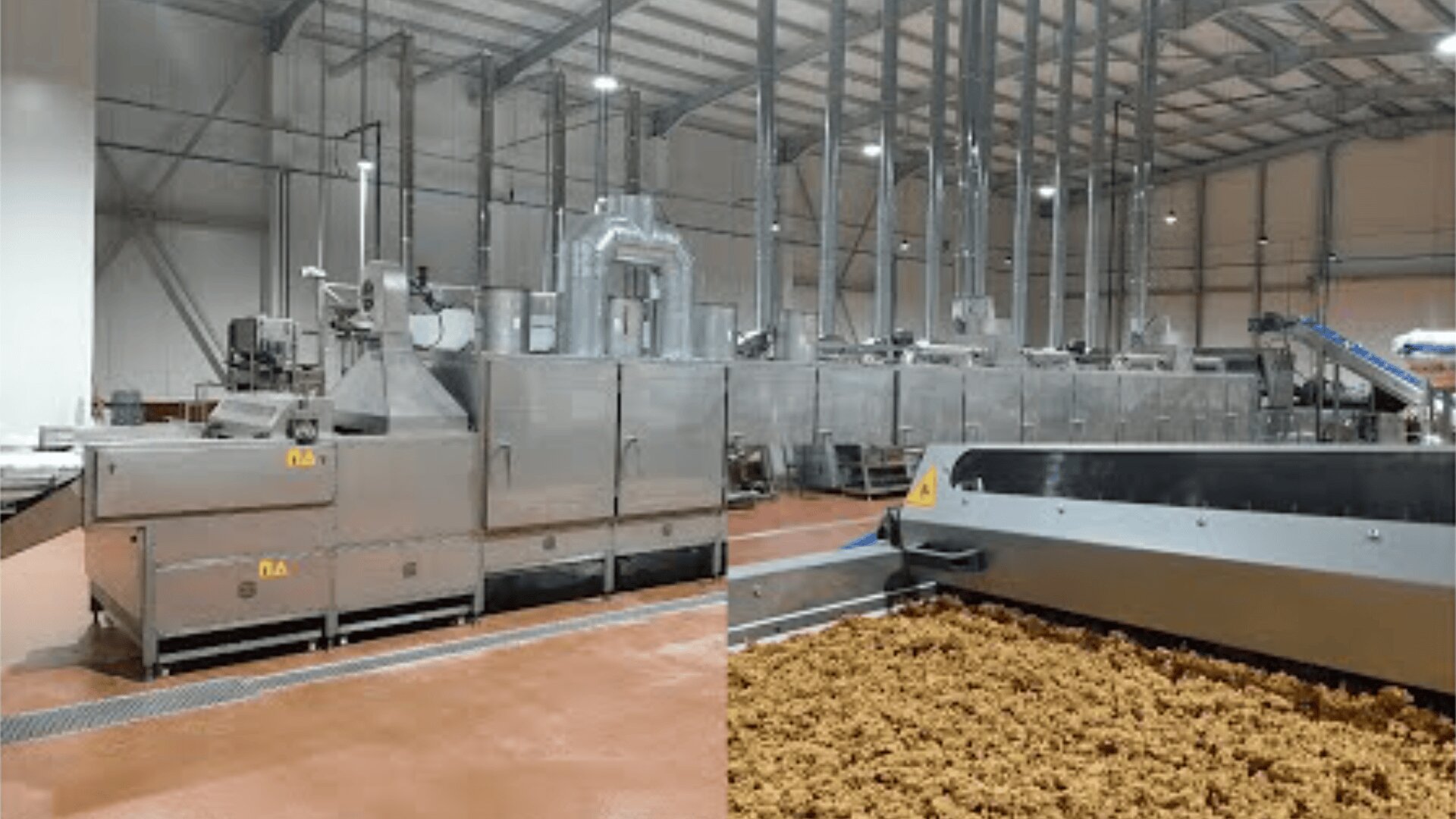 Large industrial food processing machinery in a factory with conveyor belts transporting a crumbly brown food product. The facility has high ceilings, metal structures and clean, reddish-brown floors.