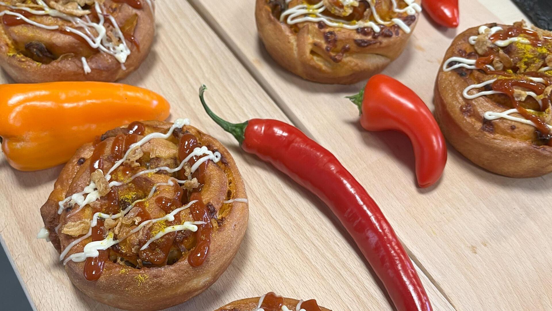 Pikante Gebäckwirbel mit weißer und orangefarbener Sauce werden neben ganzen roten und orangefarbenen Chilischoten auf einem Holzbrett angerichtet.