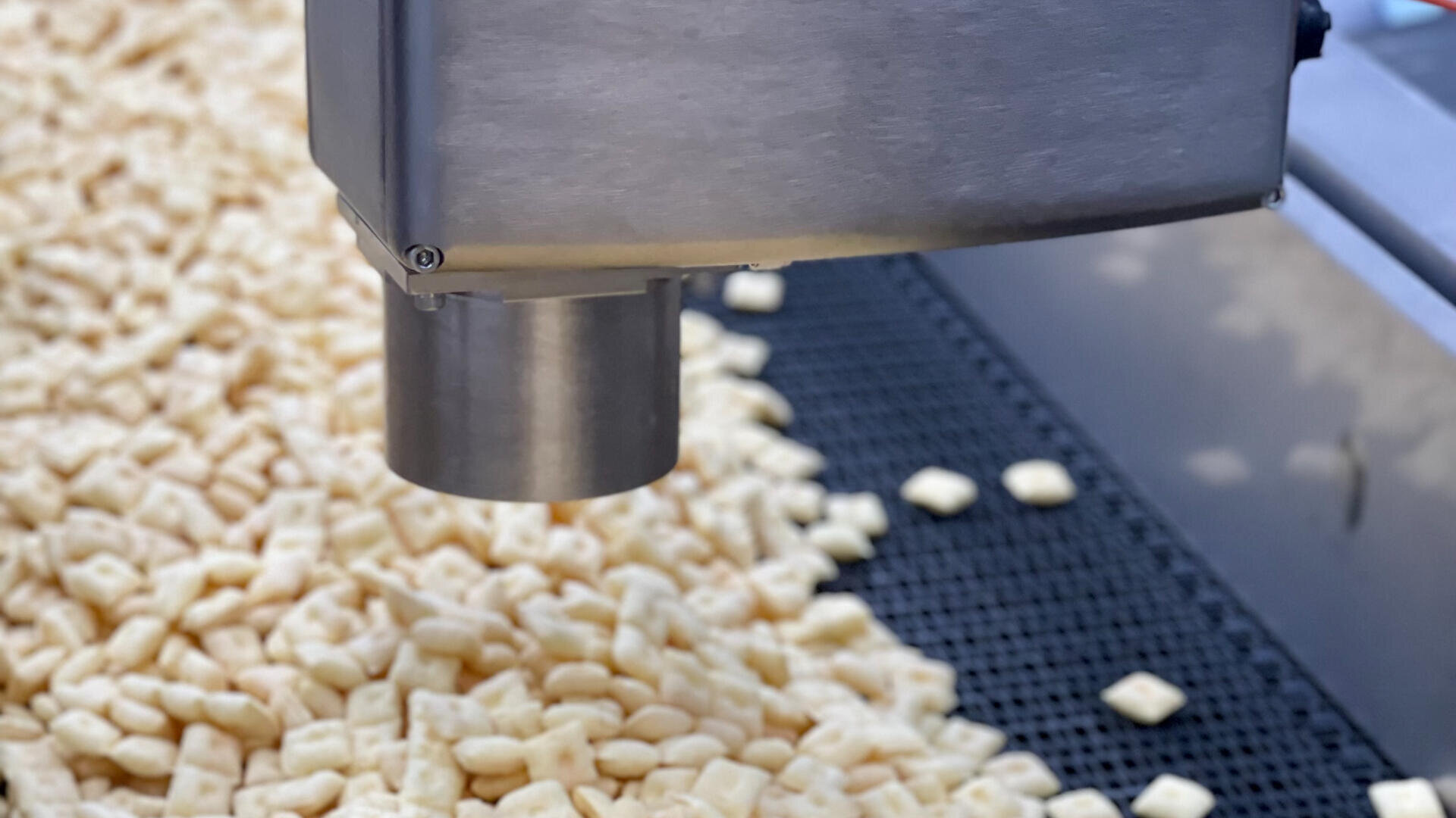 A close-up of a machine dispensing small, square pieces of food, possibly cereal, onto a conveyor belt in a food processing facility.