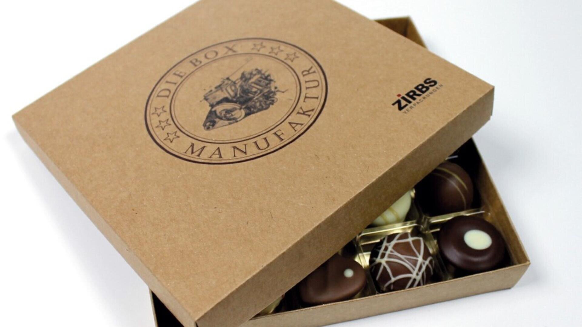 A partially open brown box labeled Die Box Manufaktur reveals assorted chocolate pralines arranged neatly inside. The box also features a small ZiRBS logo in the top right corner.