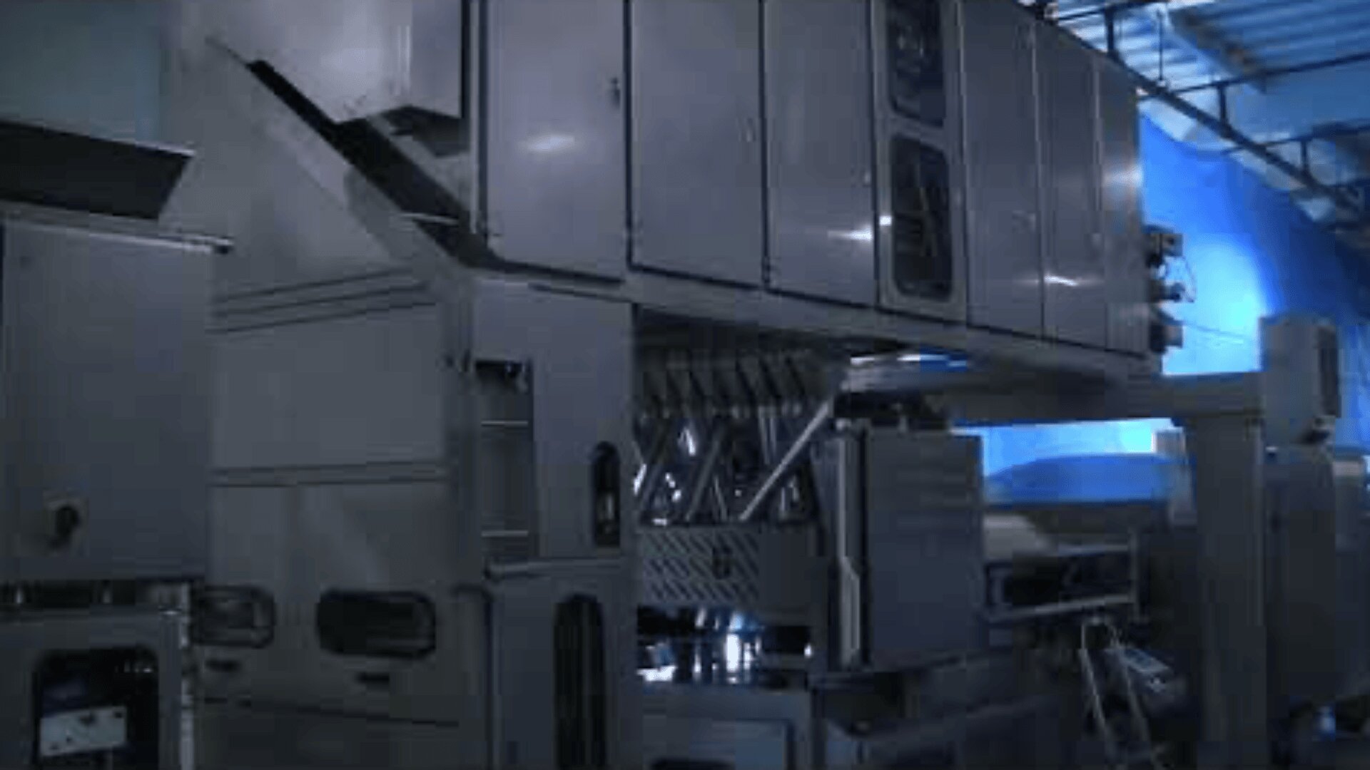Large industrial machine with metal plates, vents and pipes in a factory environment, illuminated by skylights.