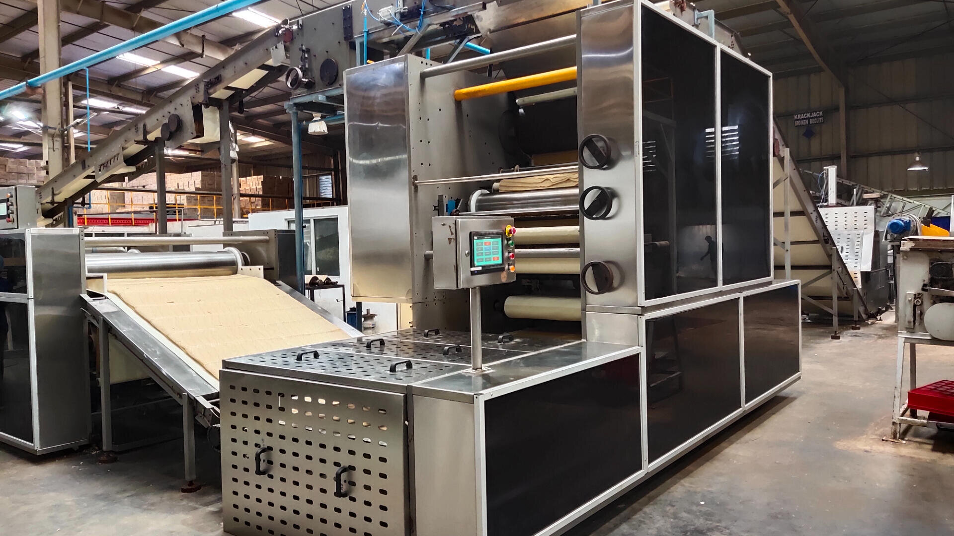 A large industrial food processing machine in a factory, with conveyor belts moving sheets of dough through stainless steel rollers and control panels nearby. The warehouse has a high ceiling and shelves in the background.