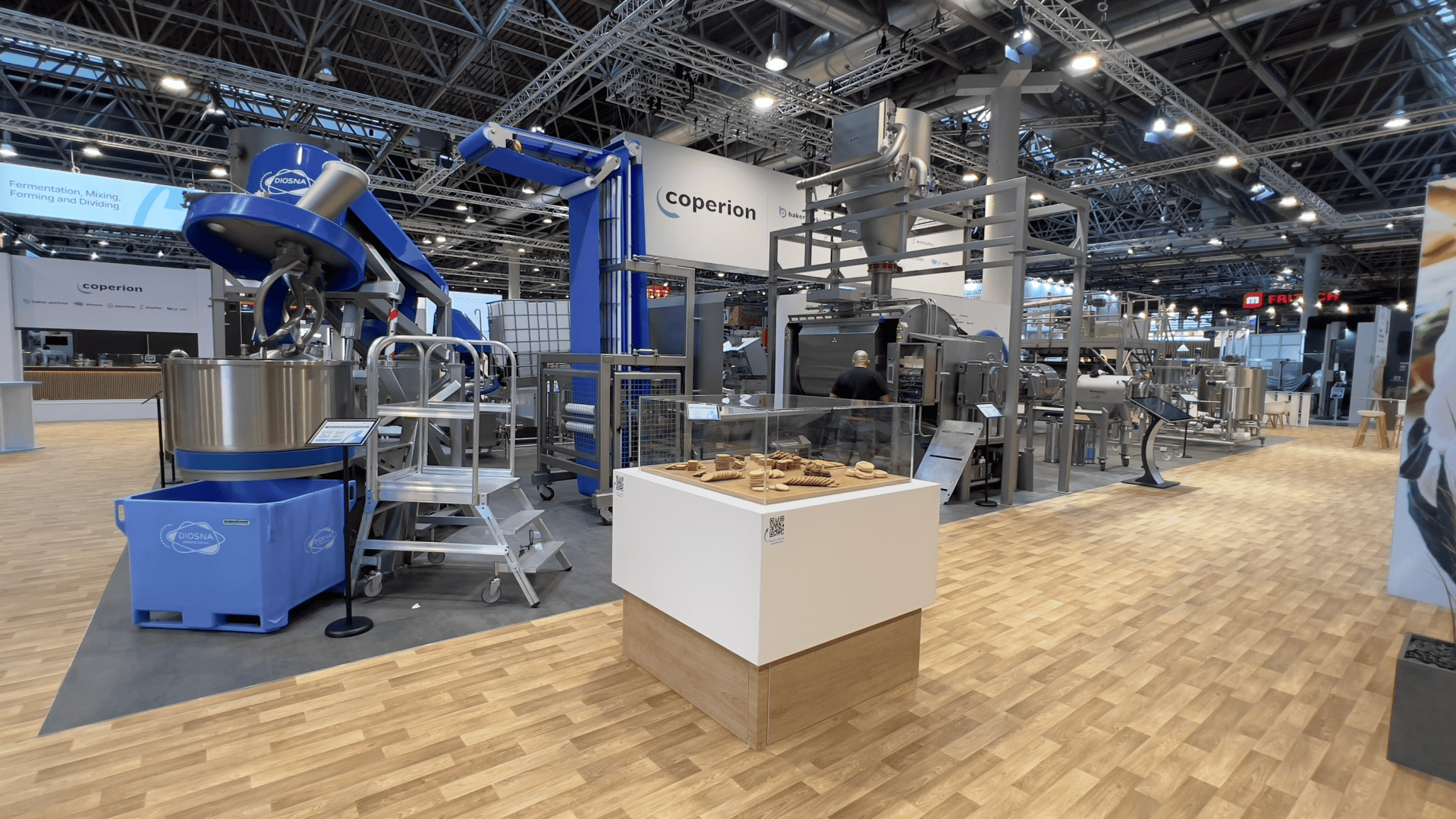 A large industrial booth at a trade show features blue and silver food processing machines, a display case with food samples, and a wooden floor. The booth is branded with Coperion signs.