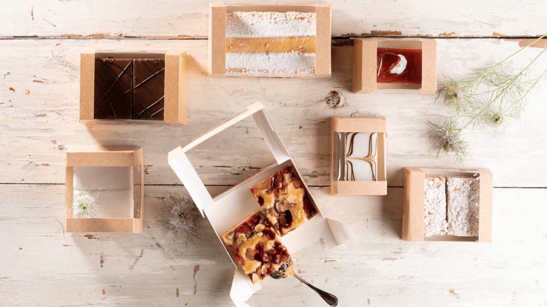 Sieben verschiedene Dessertriegel in Pappschachteln sind auf einer rustikalen weißen Holzfläche angeordnet, wobei eine offene Schachtel in der Mitte einen fruchtigen Riegel und einen Löffel zeigt. Zarte Blumenzweige sind rundherum verstreut.