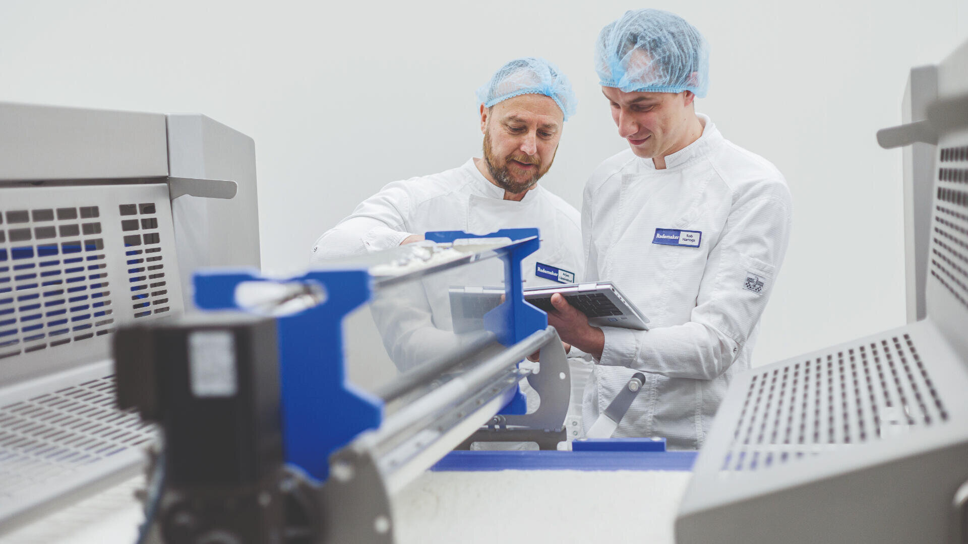 Two workers in white uniforms and blue hairnets stand next to industrial food processing equipment, reviewing notes and inspecting the machinery in a clean, bright facility.