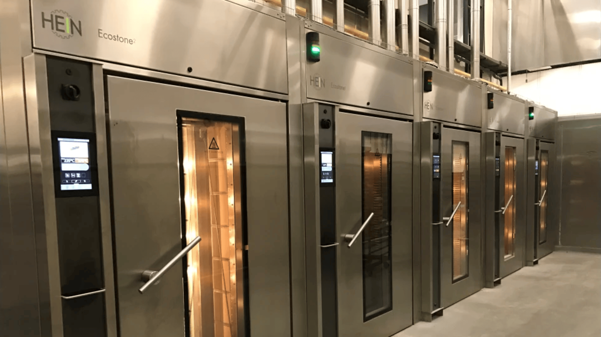 Eine Reihe großer Industrieöfen aus rostfreiem Stahl mit Glastüren und Schalttafeln, die innen beleuchtet sind, stehen in einer Großküche oder Bäckerei.