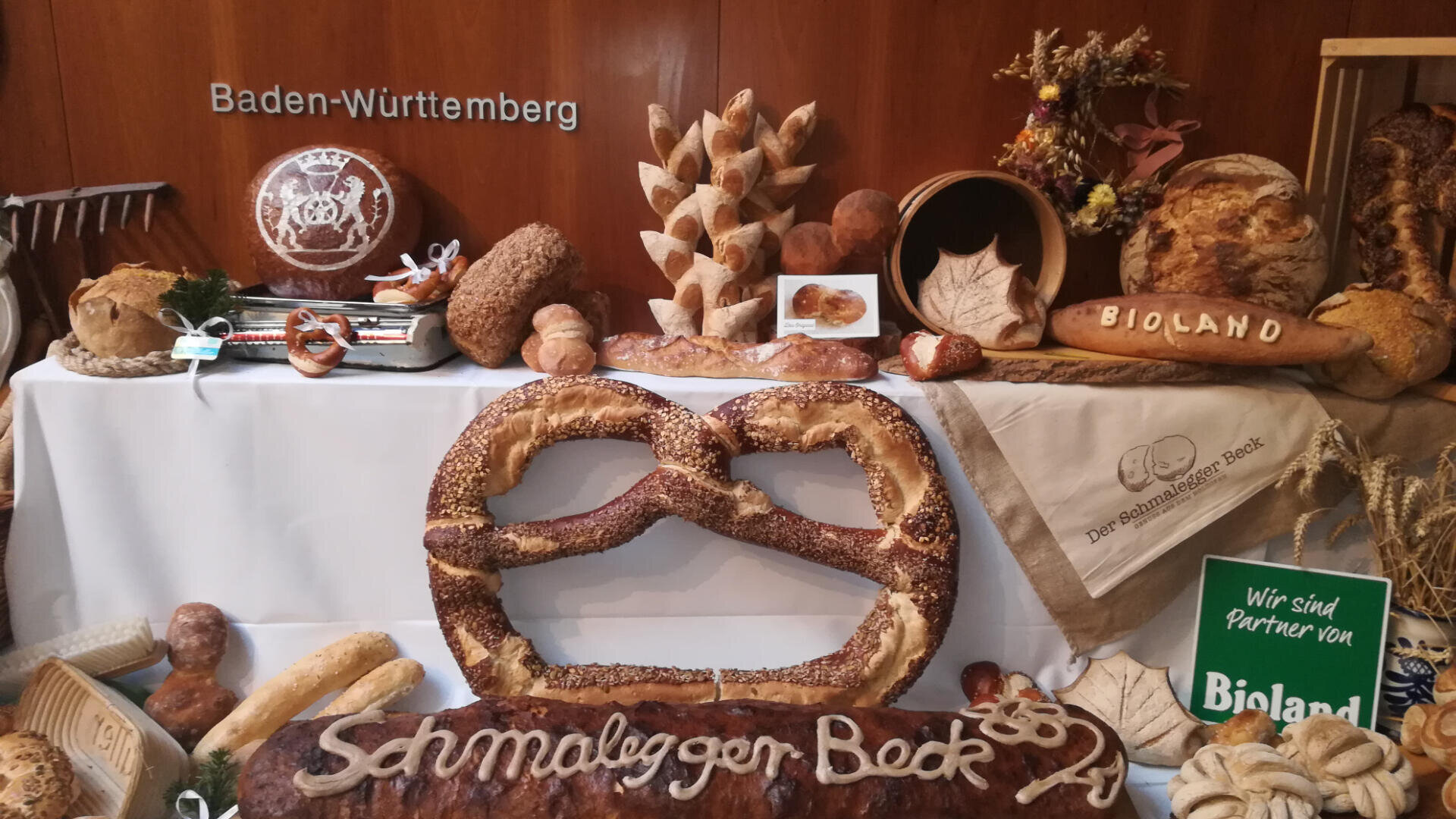 Eine Auslage mit verschiedenen handwerklich hergestellten Broten und Brezeln, darunter eine große Brezel und Brote, auf denen "Schmalzeger Back" steht. Die Einrichtung ist mit Körnern und Schildern mit der Aufschrift "Baden-Württemberg" und "Bioland" geschmückt.