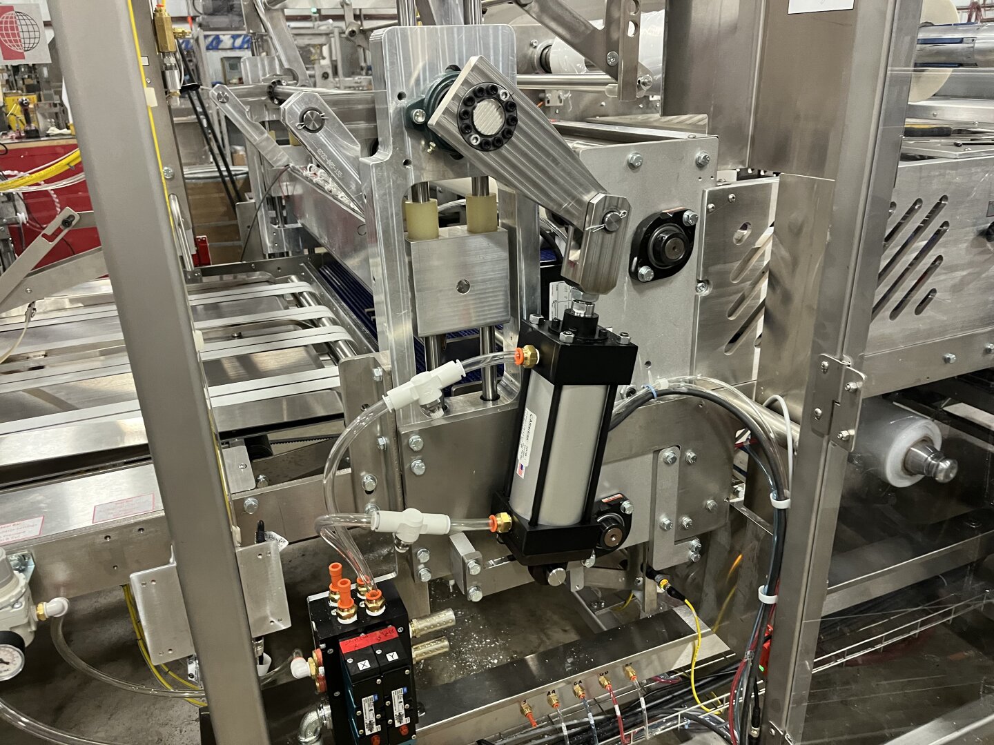 Close-up of industrial machinery with metal components, pneumatic cylinder, wires, and tubes, likely part of an automated assembly or packaging system in a factory setting.
