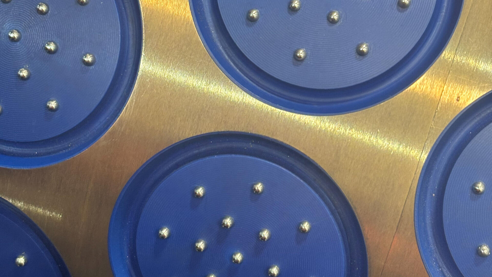 Close-up of a metallic surface with blue circular sections, each containing multiple small silver balls arranged in different patterns. The image shows a geometric and industrial design.