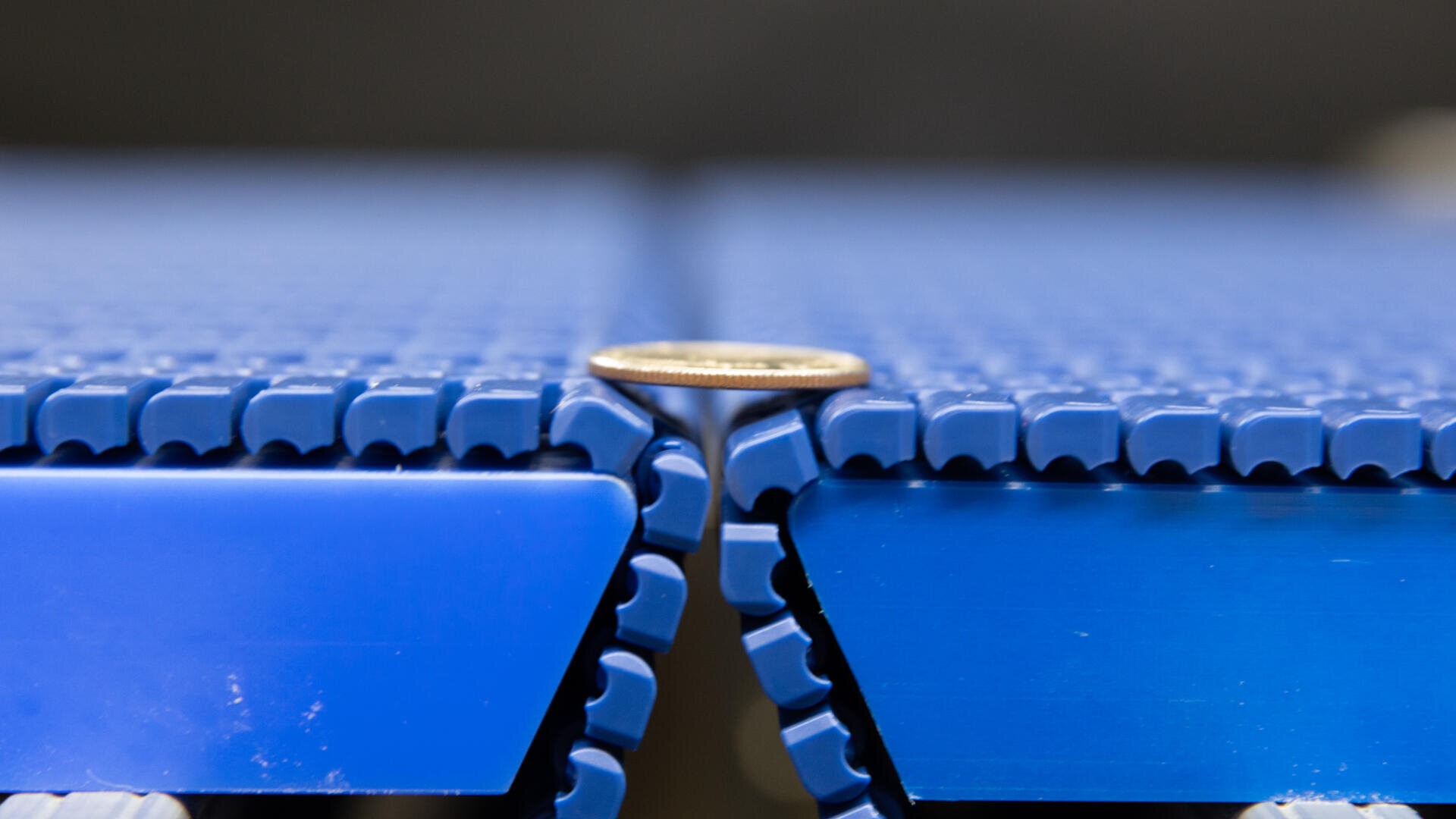 A close-up of a coin standing on its edge between two interlocking blue conveyor belts, showing the narrow gap alignment.