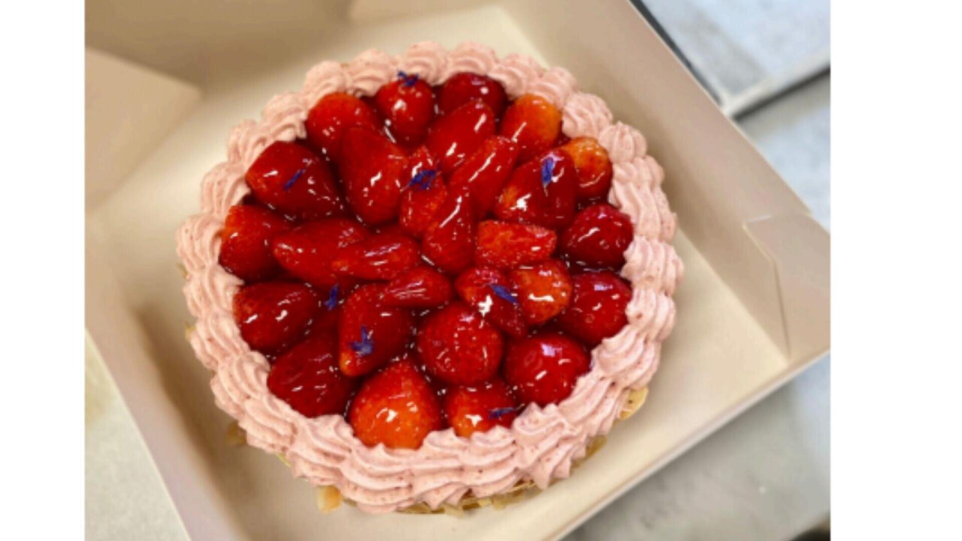 A round cake topped with glossy strawberries and decorated with pink whipped cream swirls along the edge, placed inside a white box.