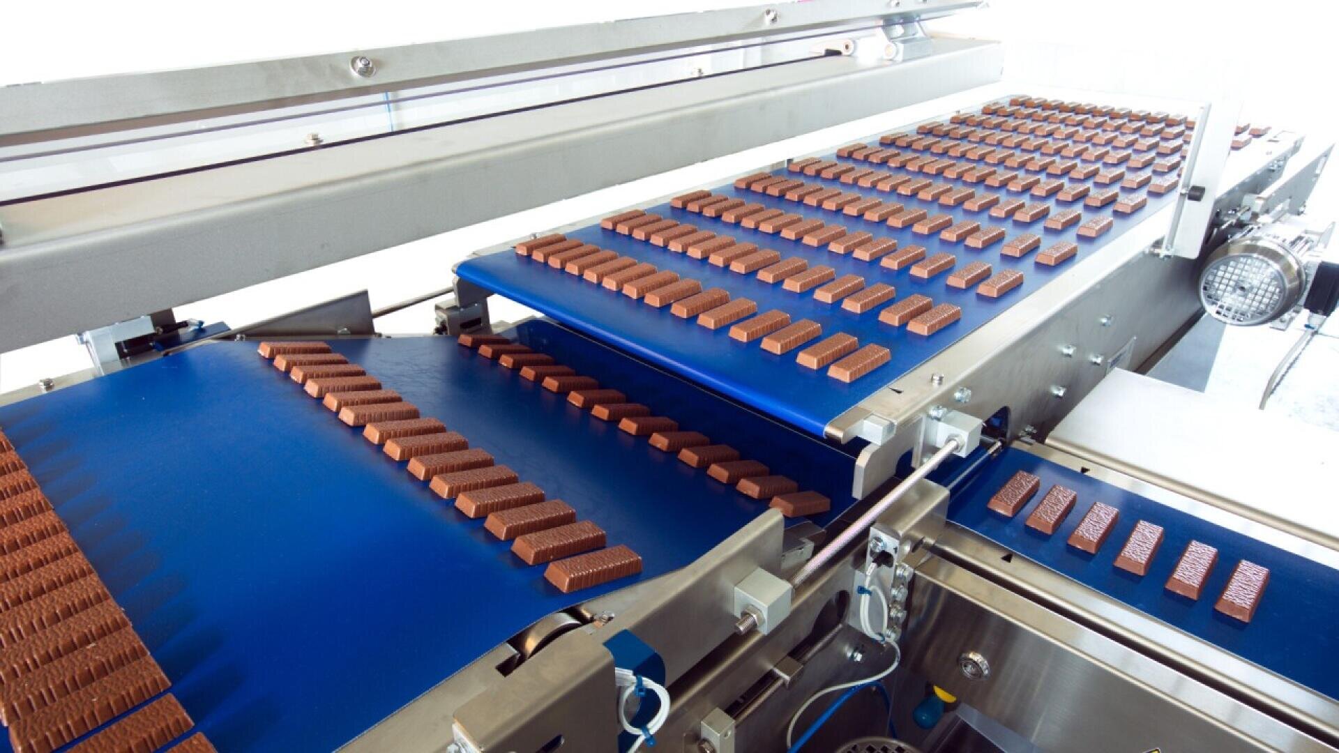 Reihen von Schokoladentafeln bewegen sich auf blauen Förderbändern in einer Fabrik, die Teil einer automatisierten Produktionslinie zur Verarbeitung und Verpackung von Schokolade ist.