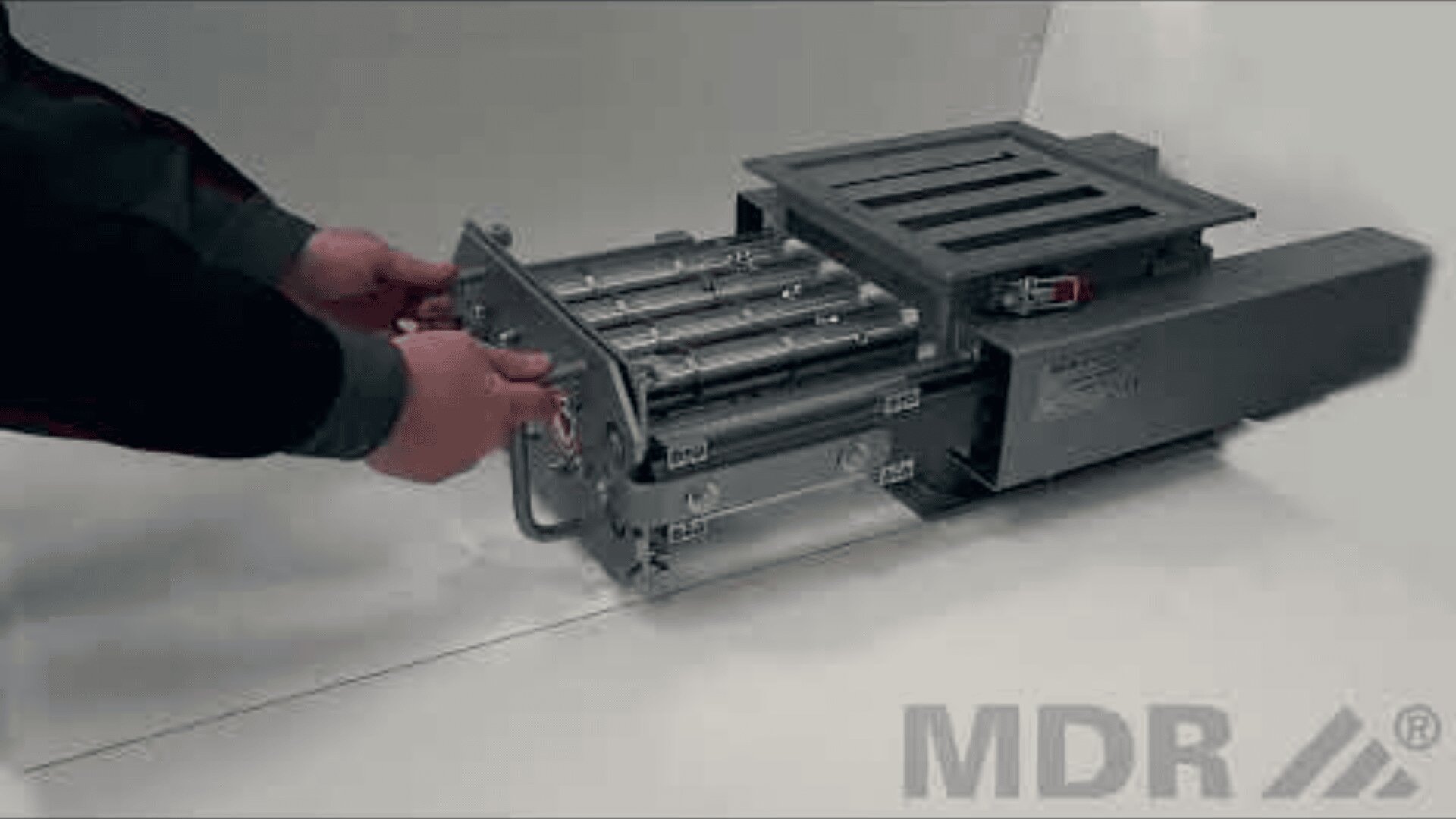 A person adjusts or checks an industrial metal device with rollers and a slotted grid standing on a white surface. The MDR logo can be seen in the bottom right-hand corner.