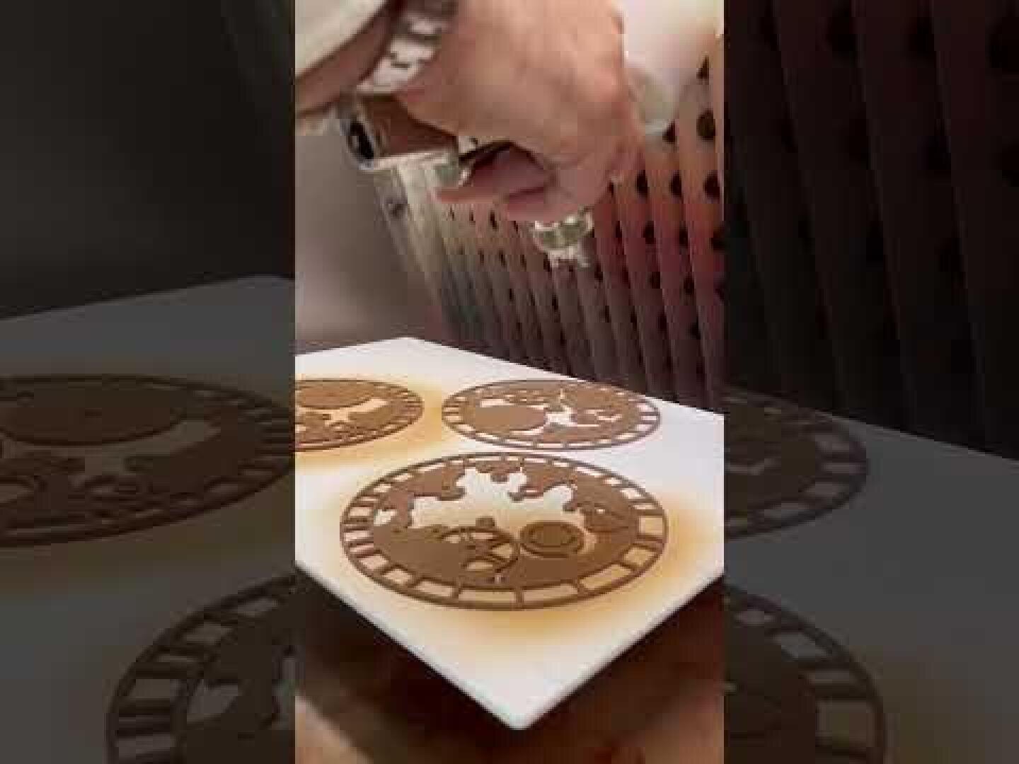 A hand holding an airbrush is spraying brown color onto three round cookies with intricate steampunk-like gear designs, placed on a white surface.