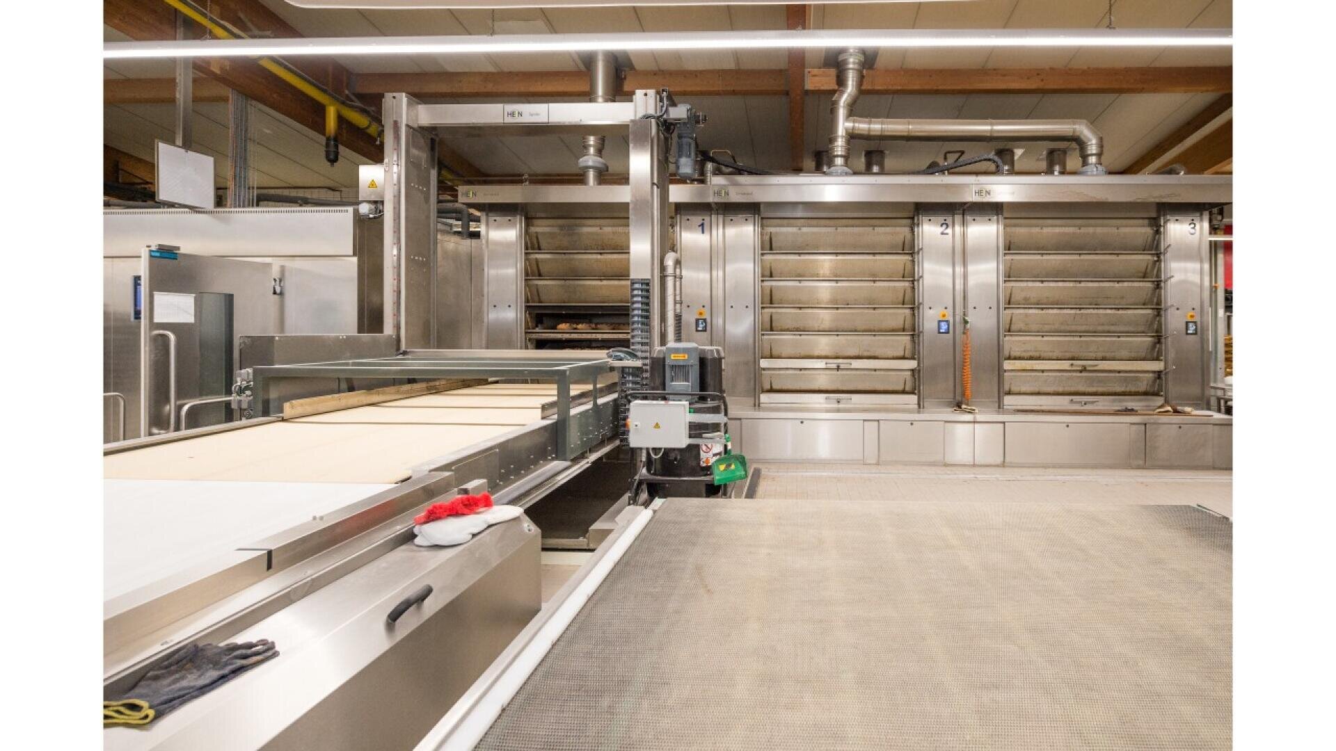 A modern industrial bakery with large stainless steel ovens, conveyor belts, and machinery. Baking trays and equipment are visible, along with red and white gloves placed on a work surface in the foreground.