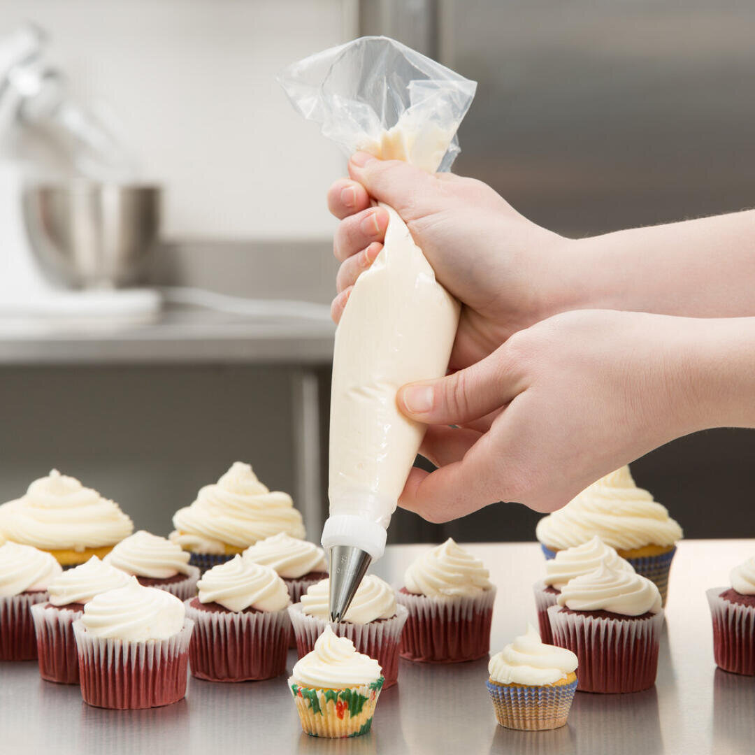 A person uses a piping bag filled with white frosting to decorate cupcakes on a metal countertop, with several already iced cupcakes in the background.