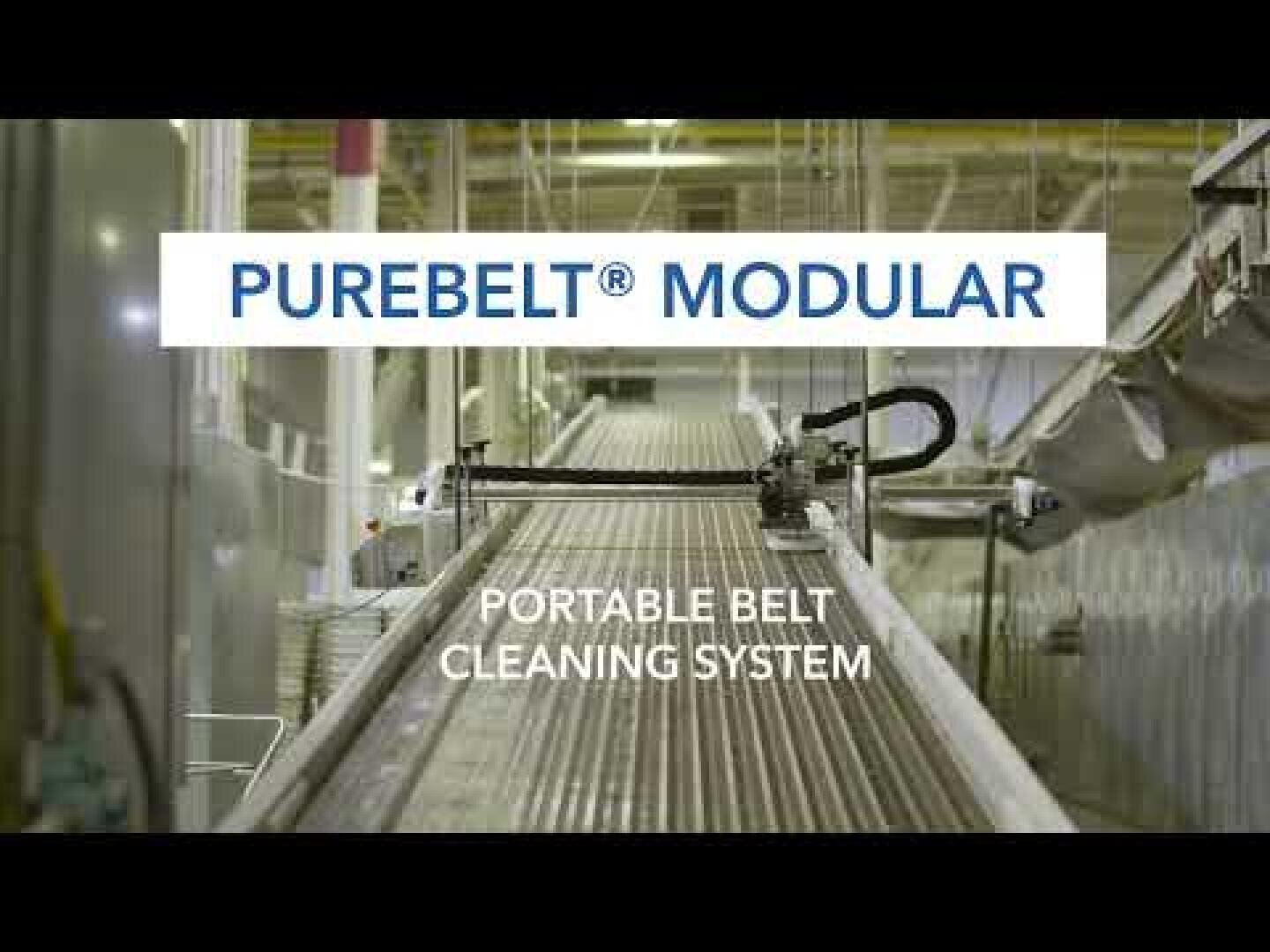 Automated machines clean a large industrial conveyor belt. Above the system in a factory facility, the text reads: PUREBELT® MODULAR AND PORTABLE BELT CLEANING SYSTEM.