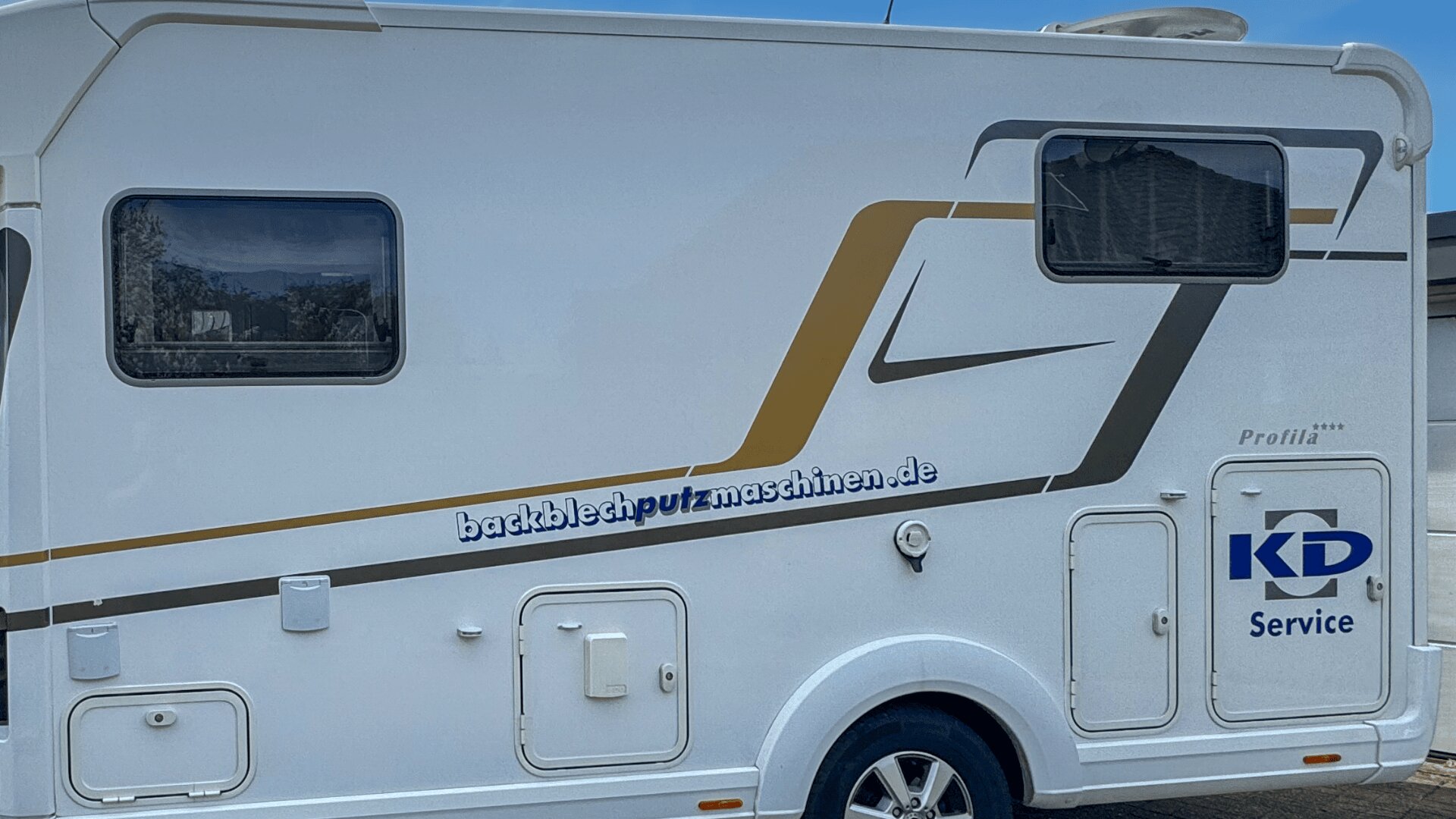 Ein weißes Wohnmobil mit goldenen und grauen Streifen, Fenstern und mehreren Außenklappen. Auf der Seite sind die Websites backblechputzmaschinen.de und KD Service aufgedruckt. Das Fahrzeug steht im Freien.
