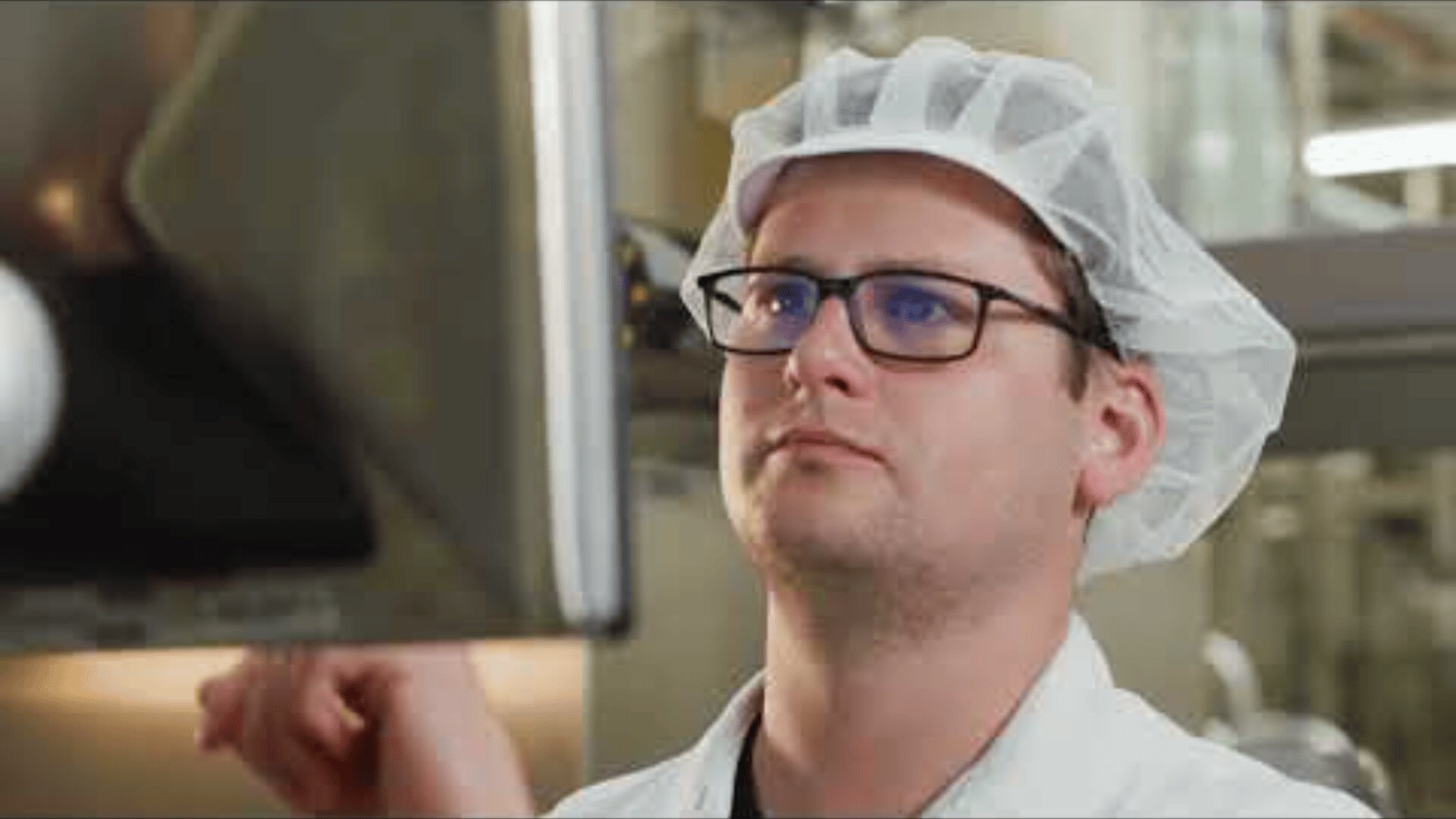 A person wearing glasses, a white hairnet, and a lab coat is focused on operating a touchscreen in a laboratory or industrial setting.