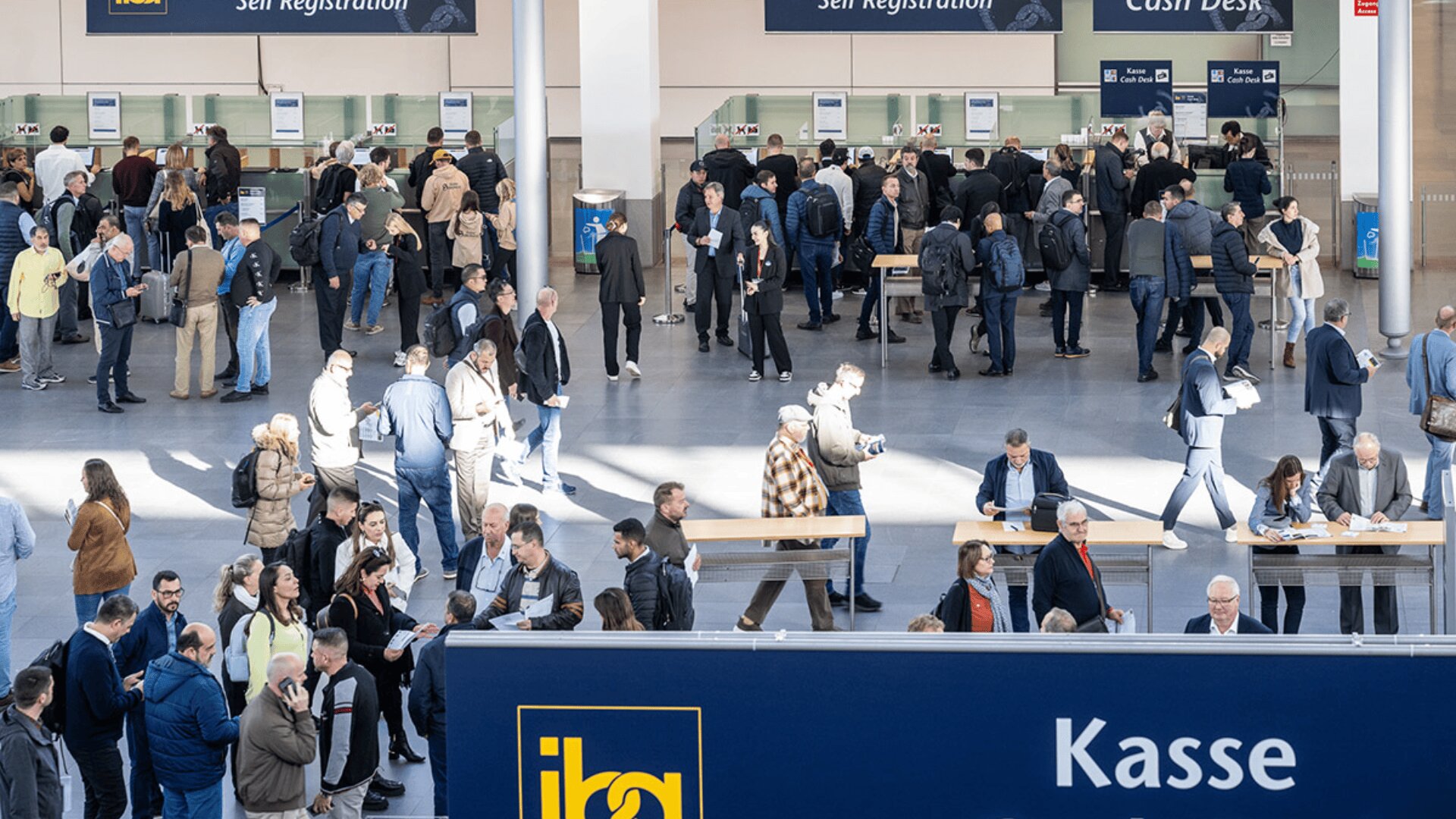 Eine belebte Kongresshalle mit vielen Menschen, die in Schlangen an den Registrierungs- und Kassenschaltern stehen, sich unterhalten und herumlaufen. Auf den Schildern steht "Self Registration", "Cash Desk" und "Kasse".