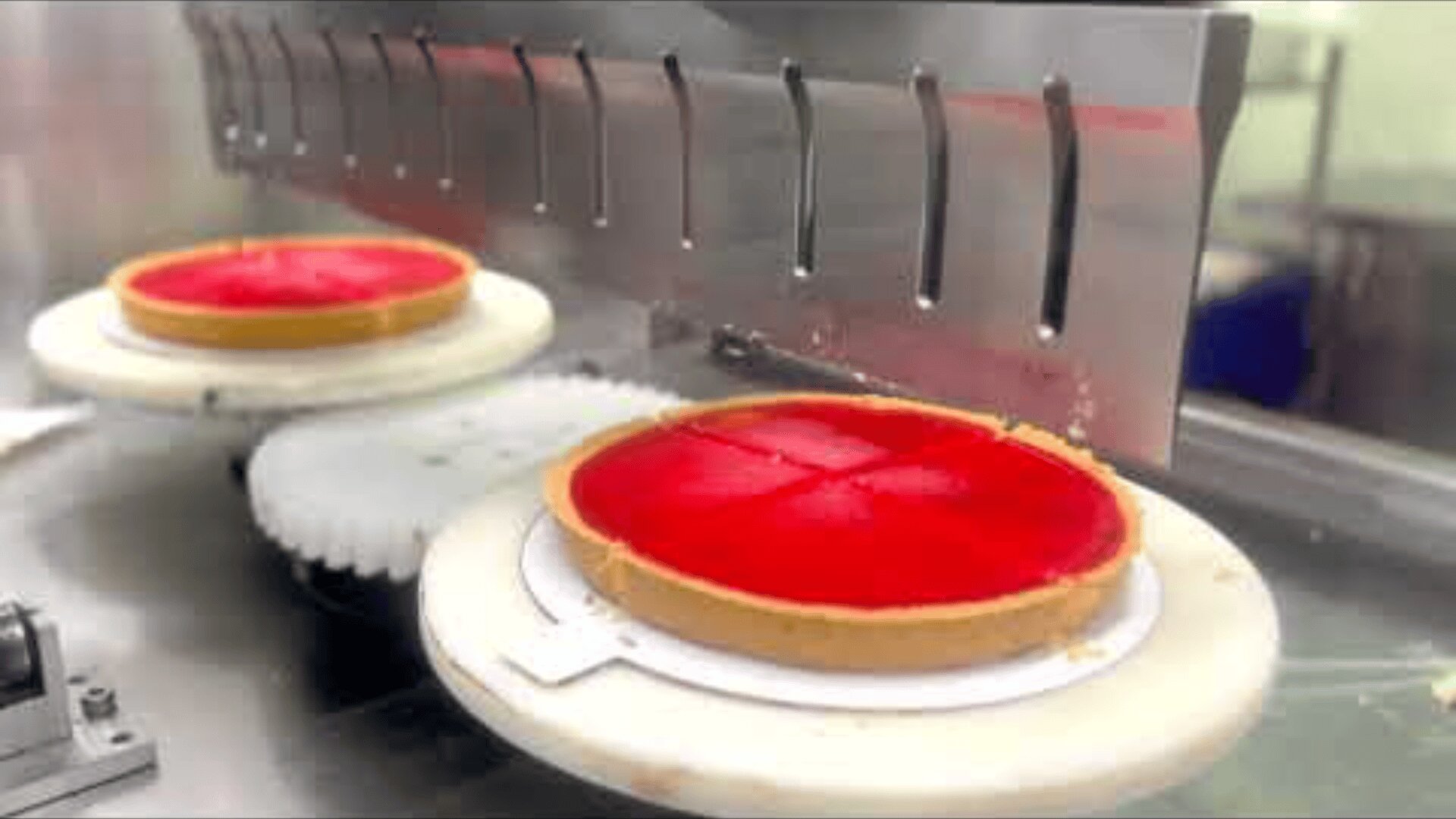 Two red cakes on round trays are placed on a conveyor belt, with one cake being cut into four pieces by a metal cutting machine.