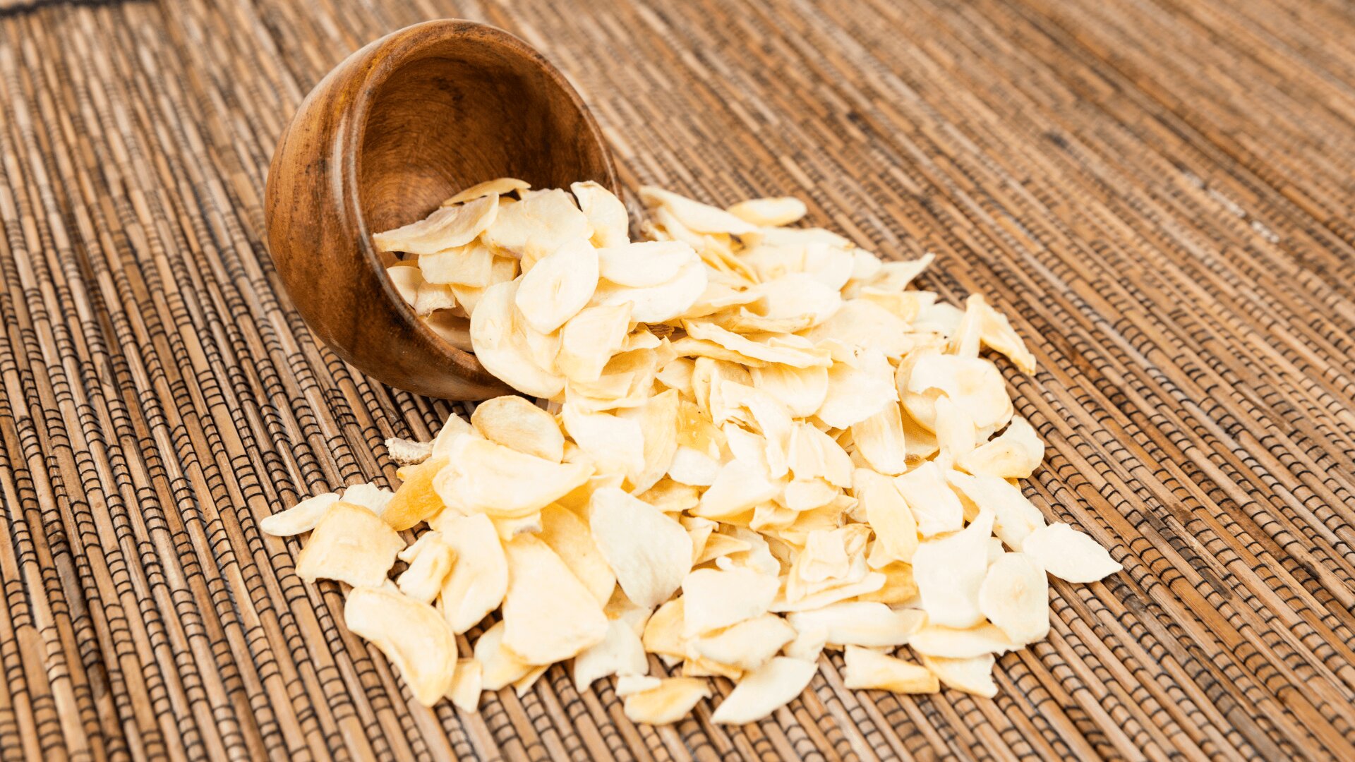 A wooden bowl tipped over, spilling dried garlic slices onto a woven bamboo mat.