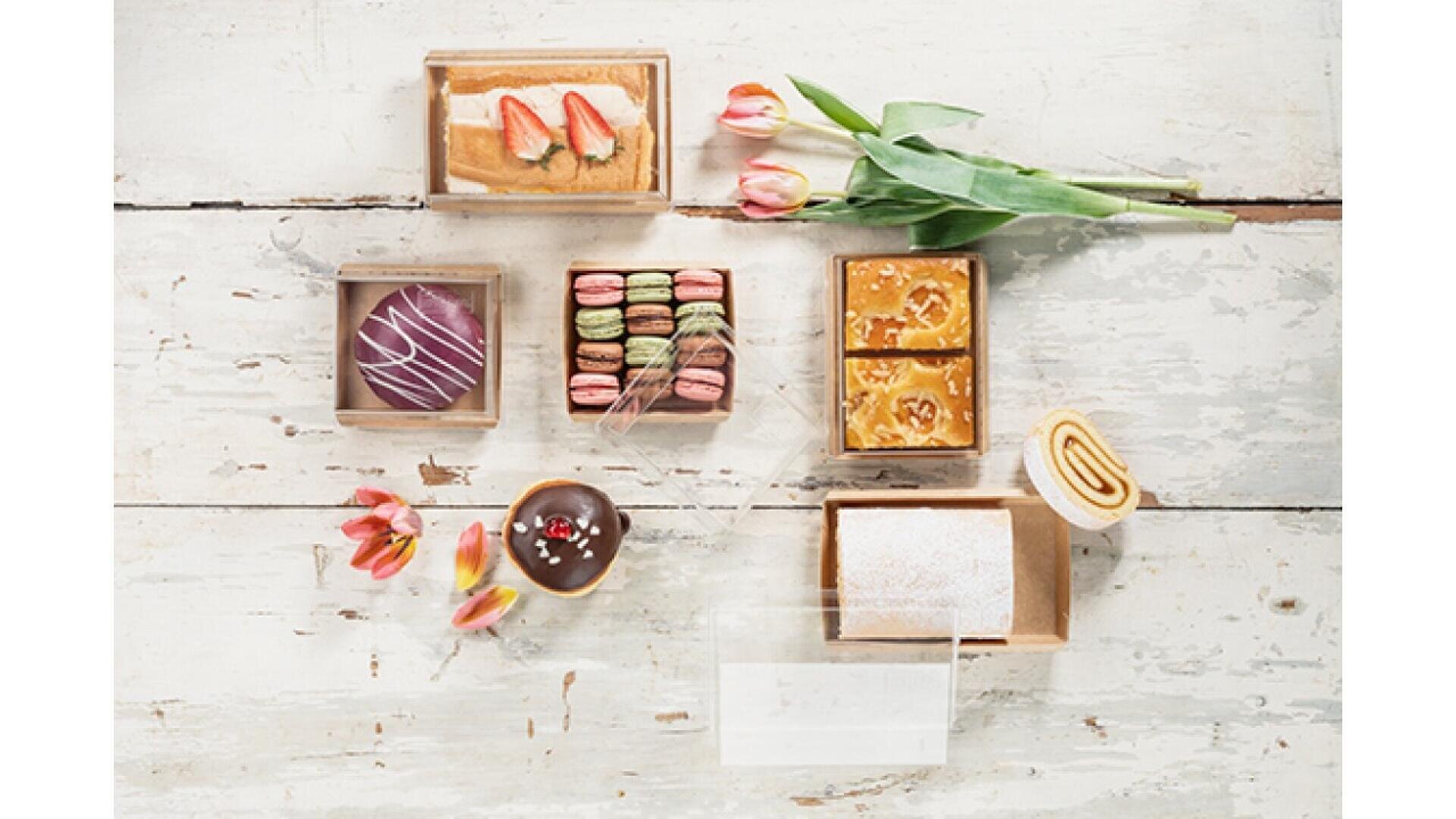 An assortment of colorful pastries, macarons, and cakes in boxes, arranged on a white rustic wooden table with a few tulips for decoration.
