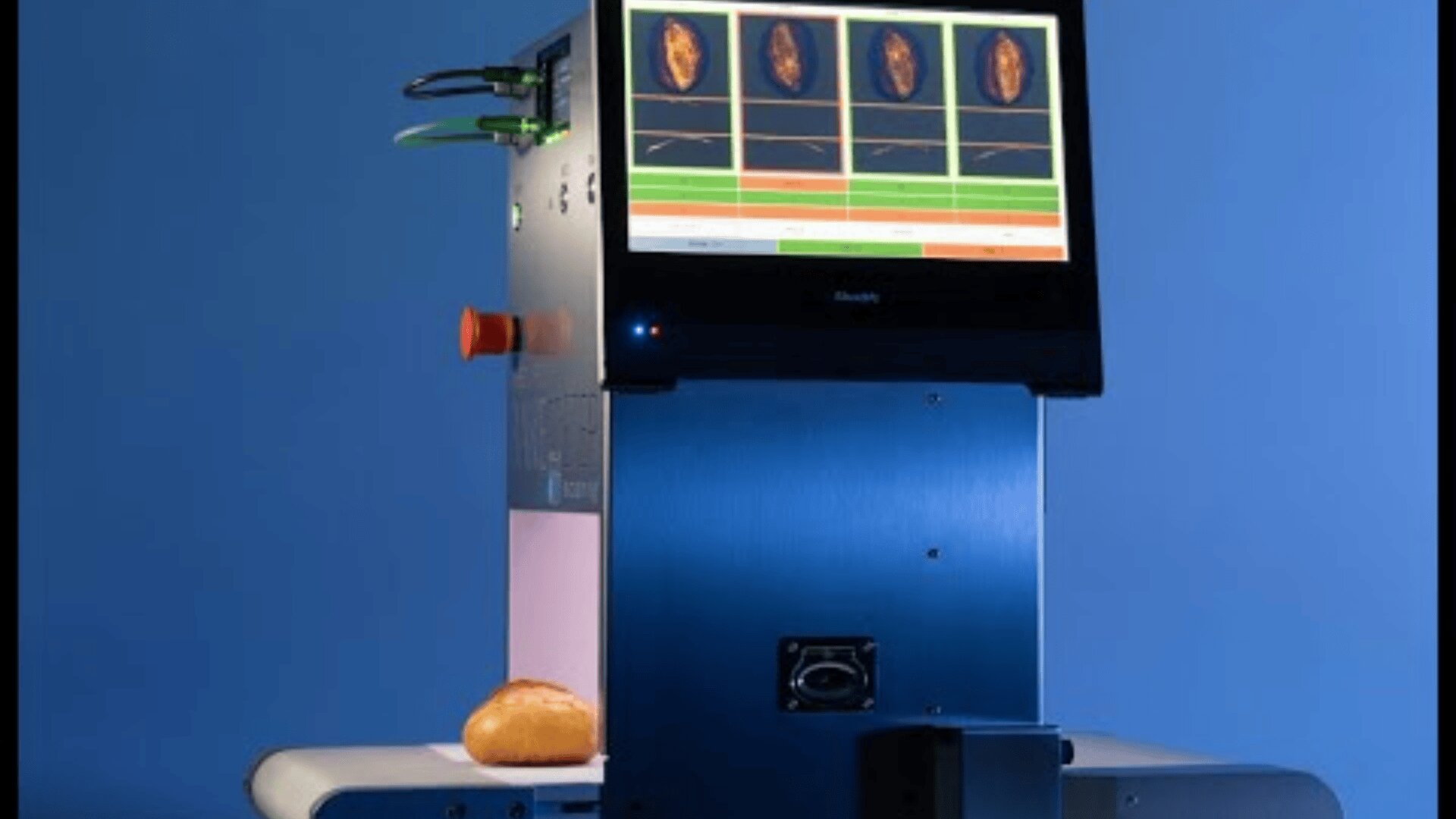 A high-tech machine with a screen displaying bread analysis scans, sits on a table with a bread roll passing through its lower section, against a blue background.