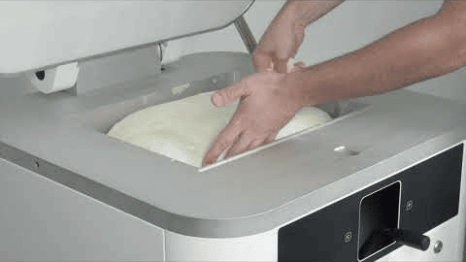 A person kneads dough in a dough kneading machine and presses the dough with both hands. The lid of the machine is open and shows the dough and the workers hands.
