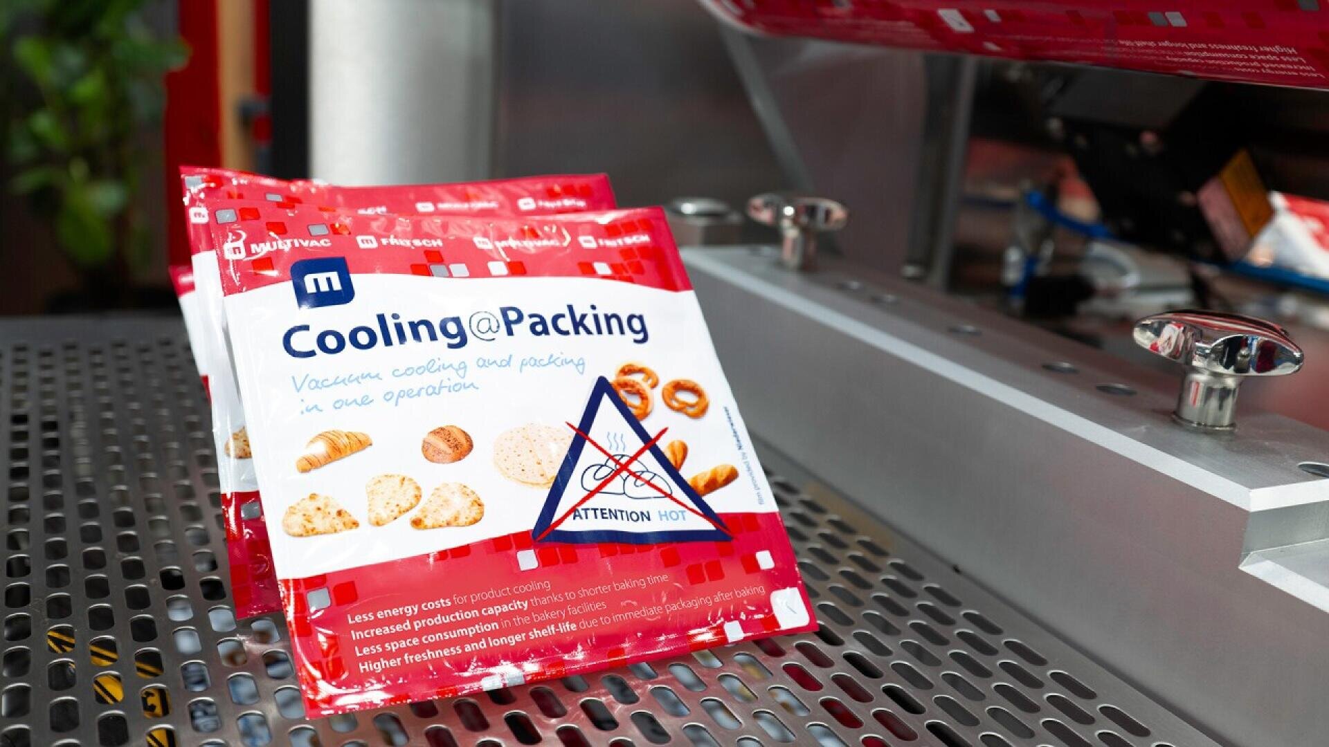 A red and white package labeled Cooling@Packing with images of baked goods sits on a metal surface near machinery. A warning label indicates Attention Hot. The packaging has various icons and text.
