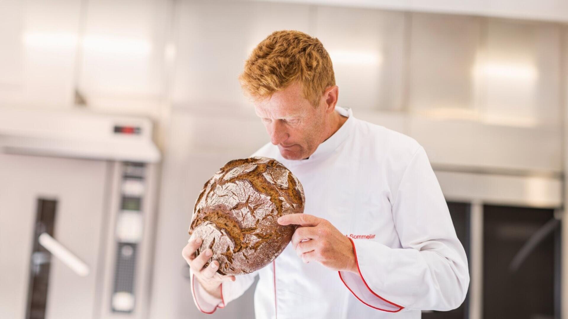 A baker in a white chefs coat holds and closely inspects a round, rustic loaf of bread in a bright, modern kitchen.